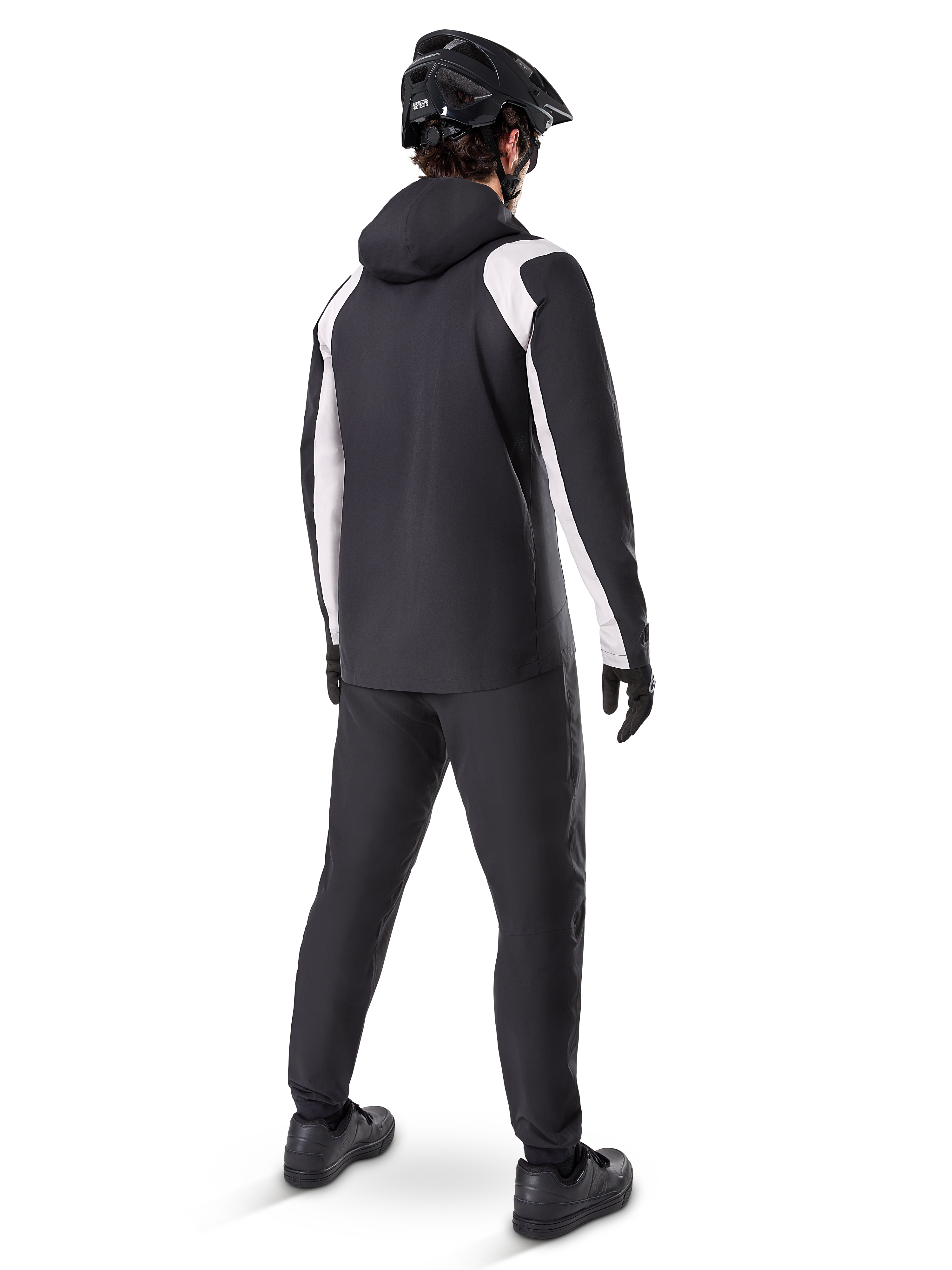 Veste de pluie Alpinestars A-Dura, vue arrière d'un cycliste portant la veste de VTT imperméable noire et blanche, pantalon de cyclisme noir assorti, casque et gants noirs, équipement technique pour le VTT par conditions humides