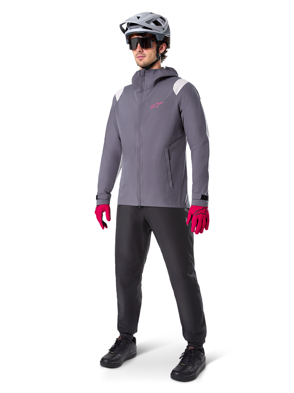 Male model wearing Alpinestars A-Dura Rain Jacket in Dark Titanium, gray hooded jacket with pink chest logo and light gray shoulder panels, paired with a gray MTB helmet, dark sunglasses, vibrant pink gloves, and black pants for off-road cycling.