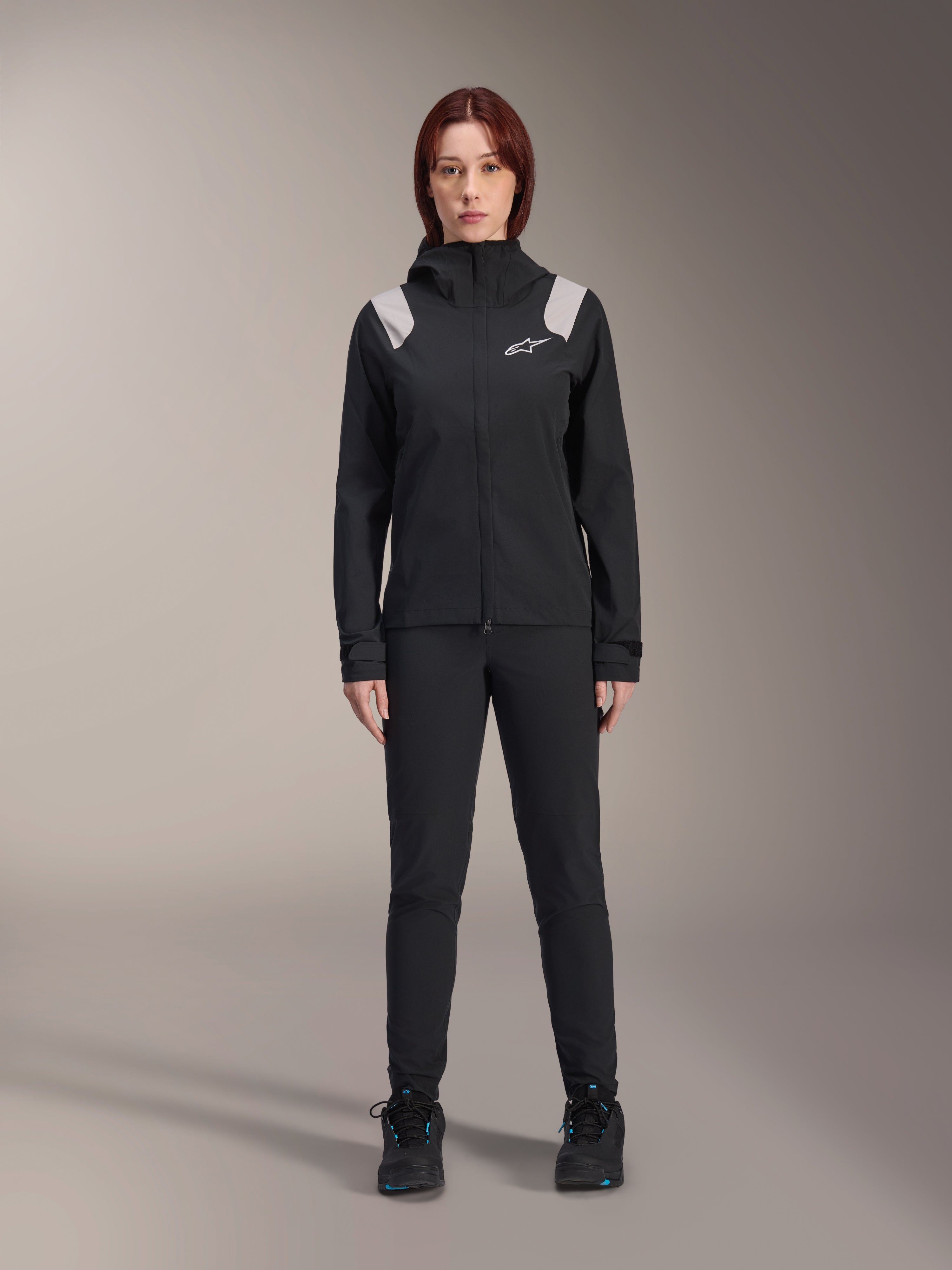 Veste de pluie Alpinestars Stella A-Dura, veste de pluie de VTT pour femme, noire avec empiècements gris clair aux épaules et logo blanc, portée avec un pantalon et des chaussures noirs assortis, dotée d'une capuche et de poignets réglables, conçue pour la pratique du VTT