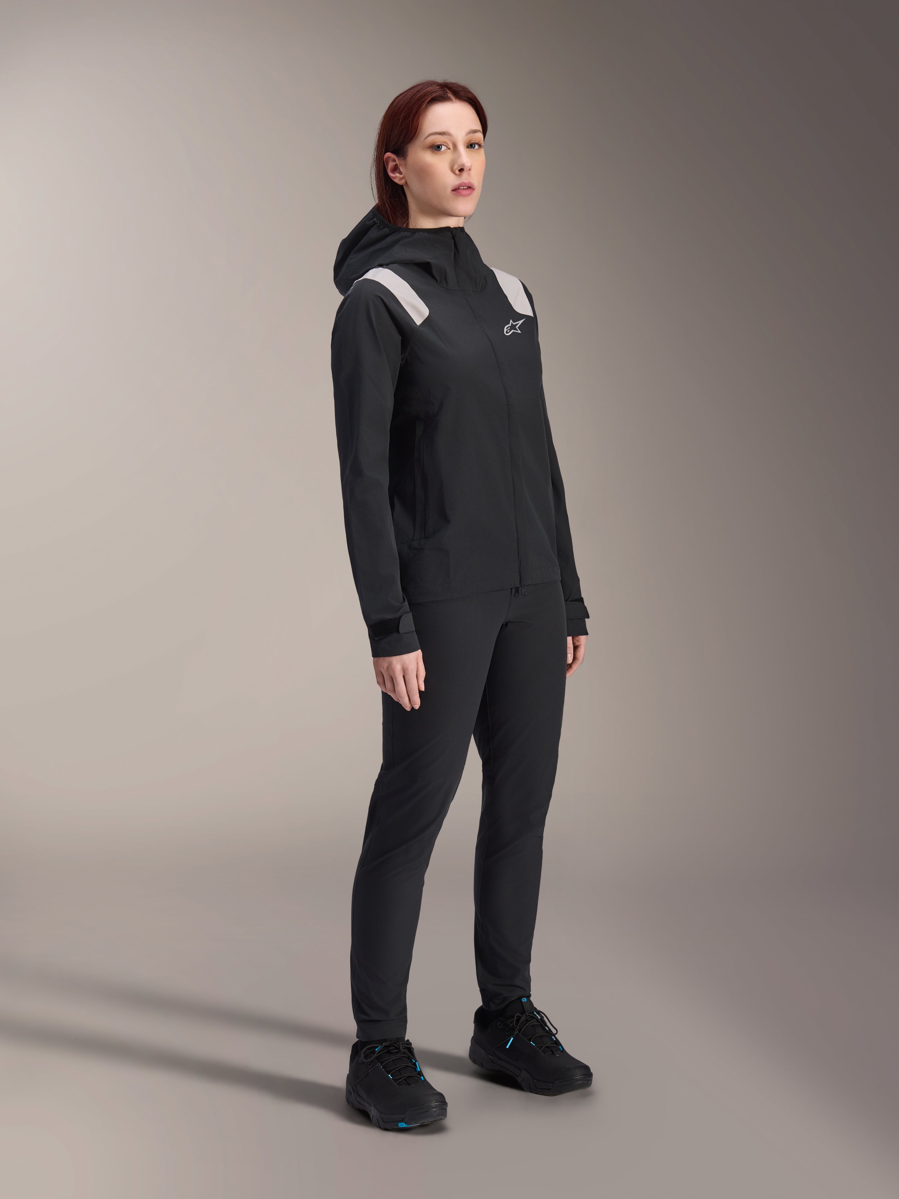 Veste de pluie Alpinestars Stella A-Dura pour femme, noir, membrane VTT imperméable 2,5 couches avec détails gris clair aux épaules, pantalon noir et chaussures de cyclisme assortis, portrait en studio pour équipement de cyclisme haute performance
