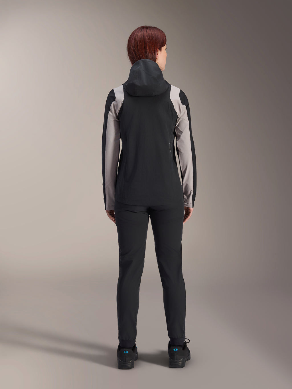Cycliste vue de dos portant la veste de pluie Alpinestars Stella A-Dura, noire avec des empiècements gris clair sur les manches et les épaules, capuche relevée, portée avec un pantalon noir et des chaussures de cyclisme, équipement technique de pluie pour le VTT