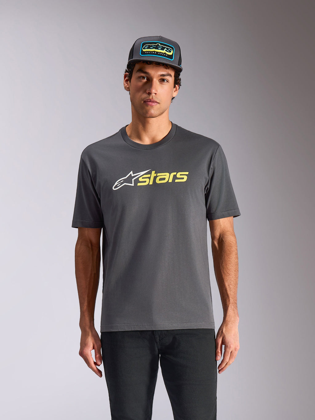 T-shirt Alpinestars Blaze 2.0 CSF, t-shirt à manches courtes, gris anthracite, logo étoile blanc et graphisme « stars » jaune fluo, porté avec une casquette trucker assortie et un pantalon noir, streetwear motorsport décontracté