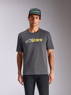 T-shirt Alpinestars Blaze 2.0 CSF, t-shirt à manches courtes, gris anthracite, logo étoile blanc et graphisme « stars » jaune fluo, porté avec une casquette trucker assortie et un pantalon noir, streetwear motorsport décontracté