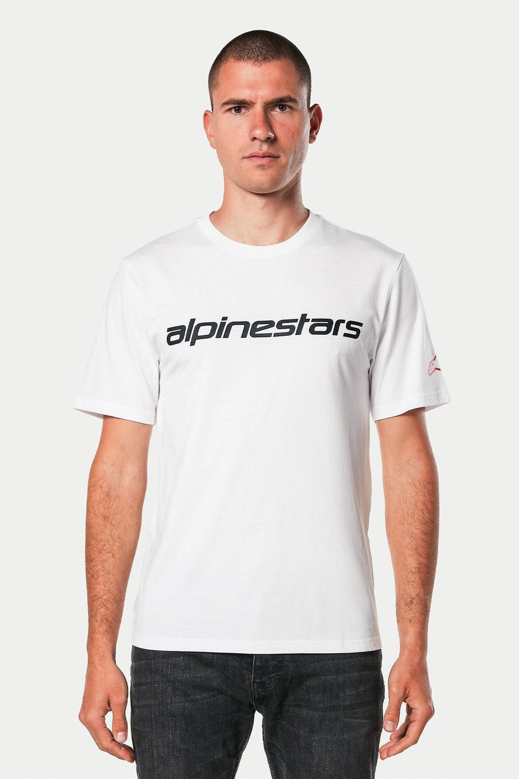 Alpinestars Linear Wordmark 2.0 CSF Tee, t-shirt à manches courtes, blanc avec mot-symbole linéaire noir sur la poitrine et logo rouge sur la manche, porté par un mannequin avec un jean foncé, streetwear décontracté inspiré du sport automobile
