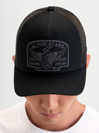 Alpinestars Casquette Trucker Heritage Patch, casquette de sport noire et grise avec visière courbée, couronne structurée et écusson brodé, portée par un modèle