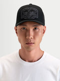 Casquette Alpinestars Casquette Trucker Heritage Patch, casquette trucker sportswear noire et grise avec visière courbée, couronne structurée et grand écusson logo brodé, portée par un modèle
