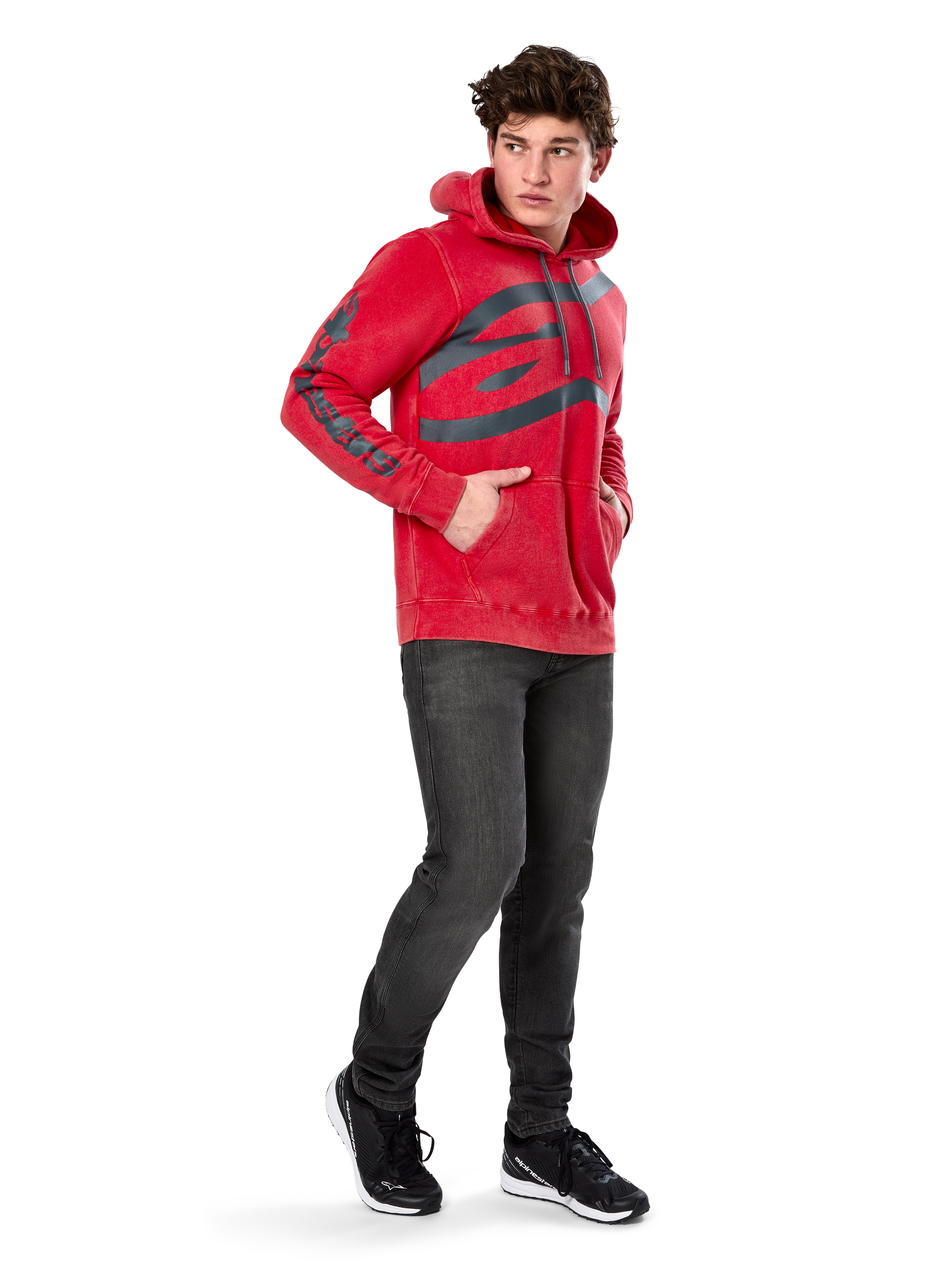 Sweat à capuche Alpinestars Unbound, polaire rouge teint en vêtement, très grand logo sombre sur la poitrine et marquage sur les manches, poche kangourou, capuche à cordon de serrage, porté par un mannequin avec un jean gris foncé et des baskets noires, streetwear décontracté inspiré du sport automobile