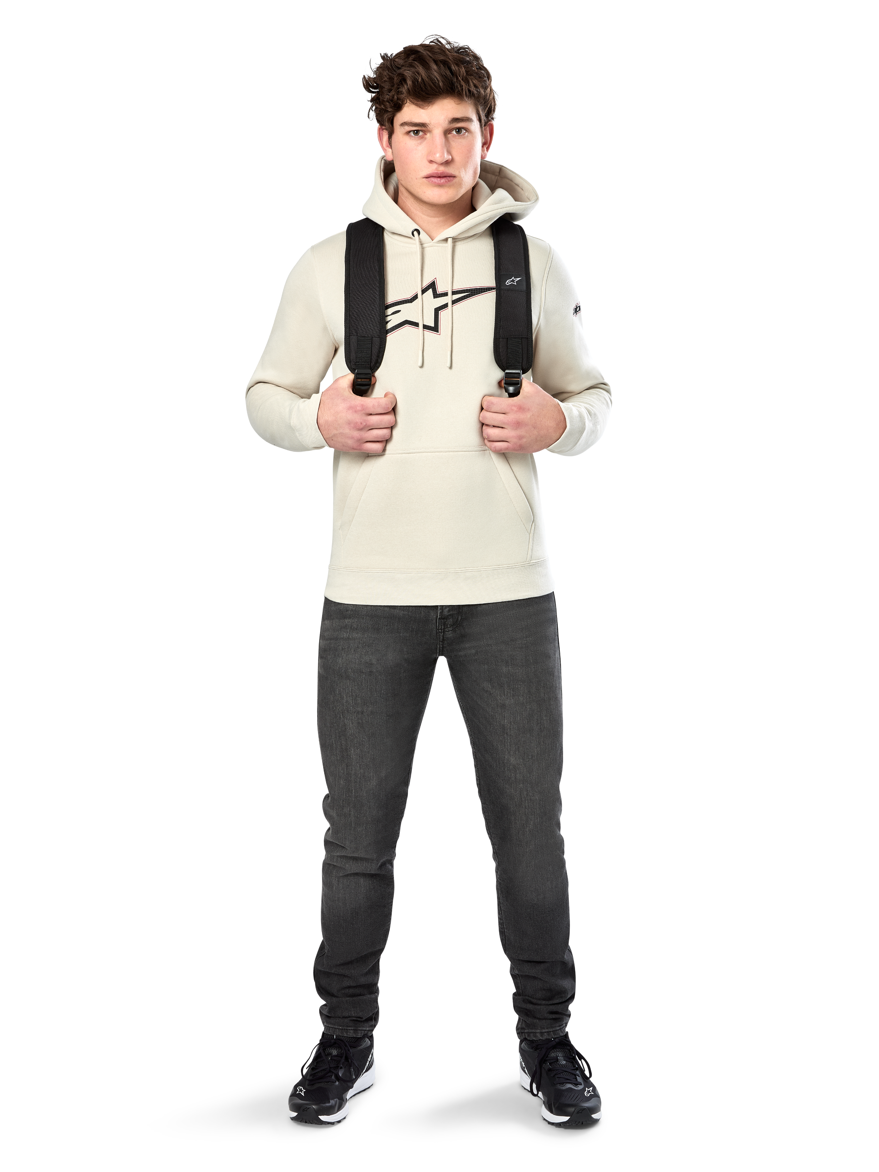 Sweat à capuche Insignia Alpinestars, streetwear décontracté, pull en polaire blanc cassé, logo aspect fibre de carbone avec contour rouge, capuche à cordon de serrage, poche kangourou, porté par un mannequin avec un sac à dos noir et un jean gris foncé