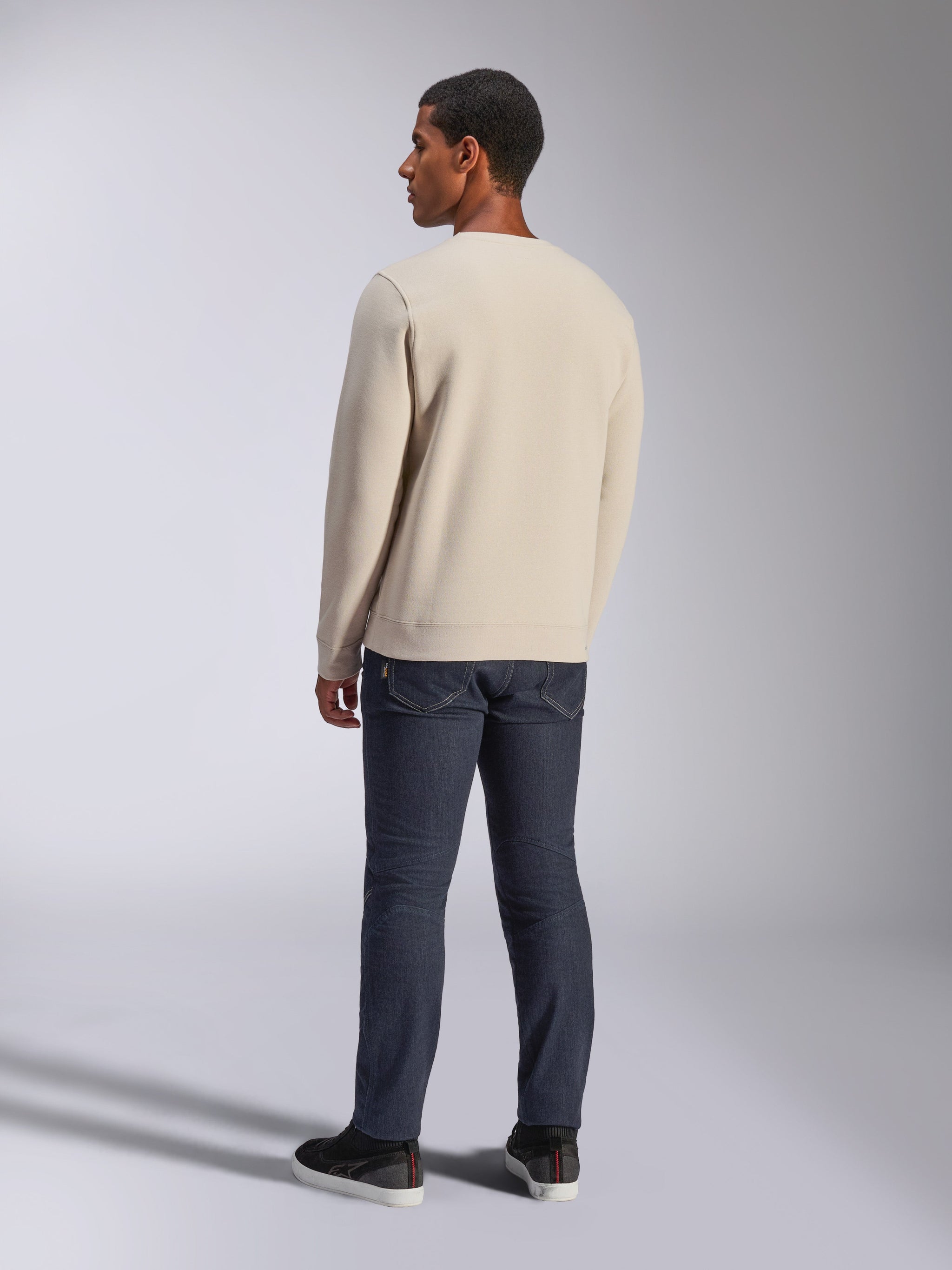 Alpinestars Positive Crew, sweatshirt à col rond blanc cassé, vue de dos sur mannequin, associé à un jean en denim foncé et des baskets noires, streetwear décontracté style motorsport, avec poignets et ourlet côtelés