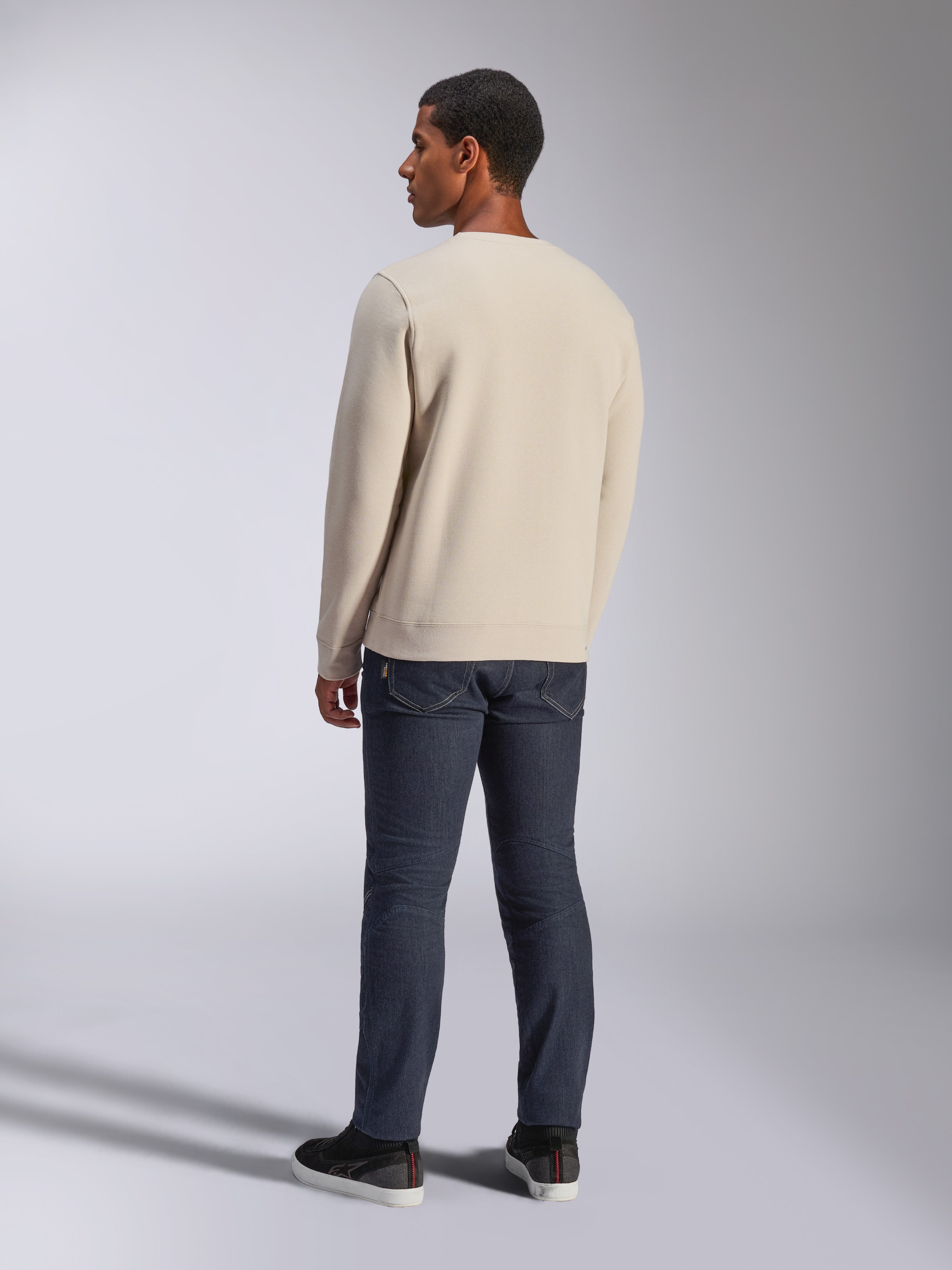 Alpinestars Positive Crew, sweatshirt à col rond blanc cassé, vue de dos sur mannequin, associé à un jean en denim foncé et des baskets noires, streetwear décontracté style motorsport, avec poignets et ourlet côtelés