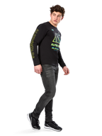 Man wearing Alpinestars Ayess CSF Long Sleeve Tee, Black, featuring yellow sleeve lettering and vibrant lime and cyan chest graphics, paired with dark grey jeans and black sneakers, casual motorsport-inspired streetwear.