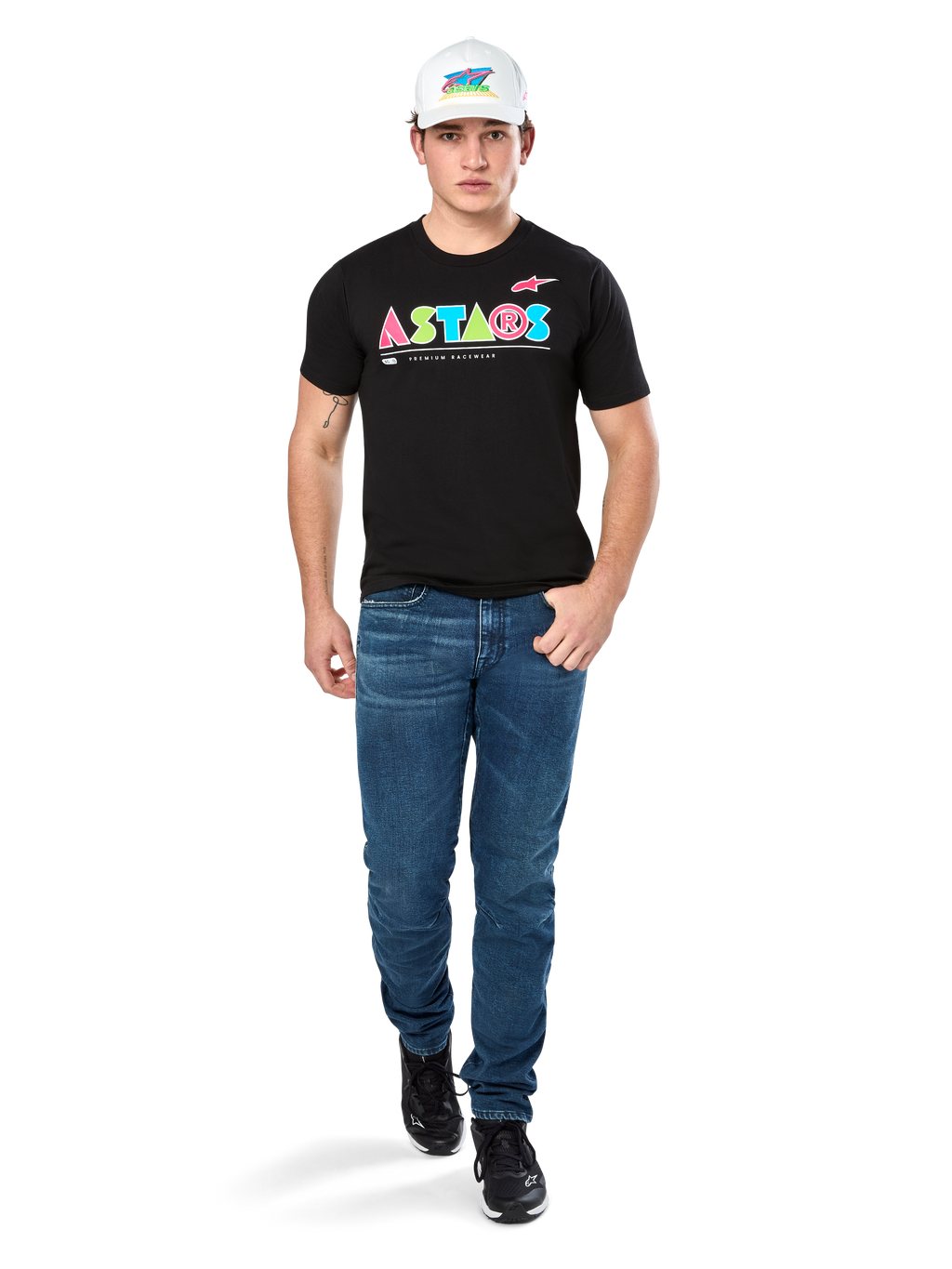 Modèle masculin portant le t-shirt Alpinestars Cereal CSF, t-shirt noir à manches courtes avec logo ASTARS géométrique coloré, associé à un jean bleu et une casquette blanche, style streetwear décontracté inspiré du sport automobile.
