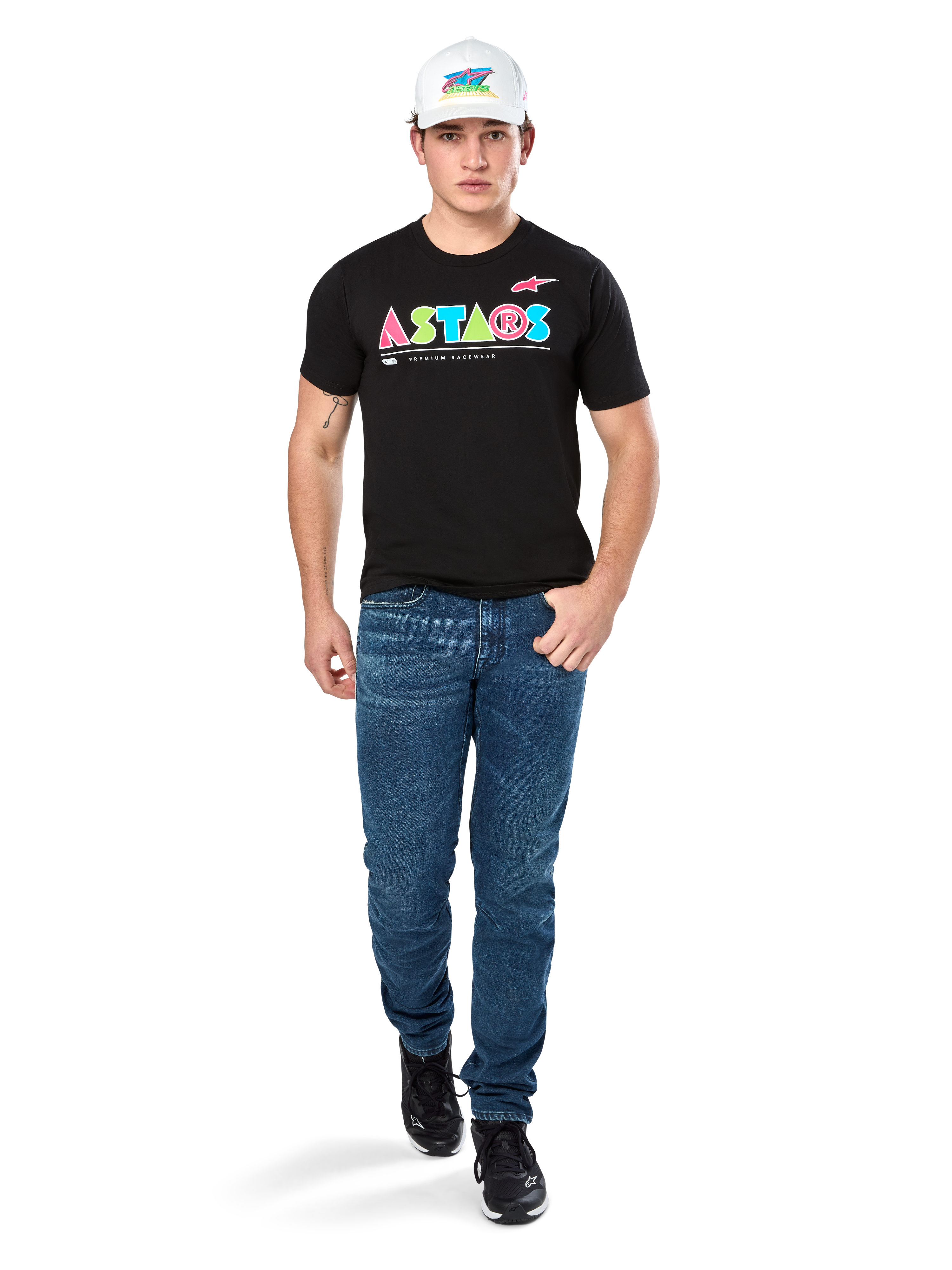 Modèle masculin portant le t-shirt Alpinestars Cereal CSF, t-shirt noir à manches courtes avec logo ASTARS géométrique coloré, associé à un jean bleu et une casquette blanche, style streetwear décontracté inspiré du sport automobile.