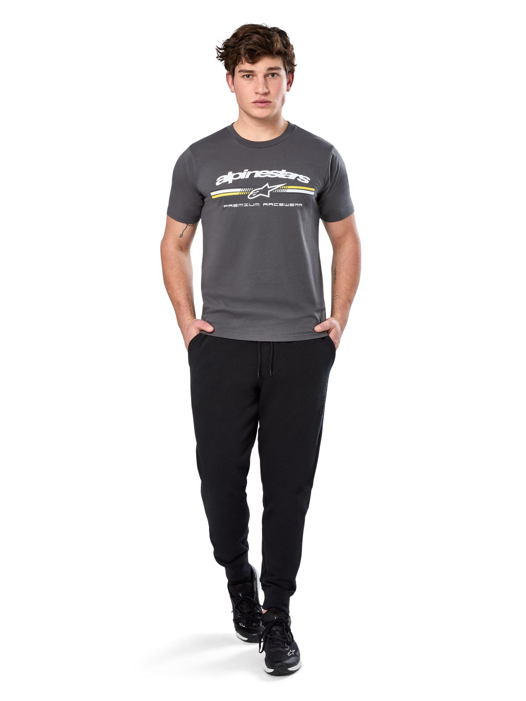 Homme portant le t-shirt à manches courtes Alpinestars Prevail CSF, gris anthracite, avec logo Alpinestars blanc et rayure tricolore aux accents jaunes, associé à un pantalon de jogging noir et des baskets, style motorsport décontracté