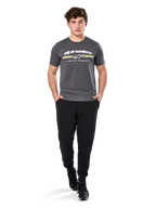 Homme portant le t-shirt à manches courtes Alpinestars Prevail CSF, gris anthracite, avec logo Alpinestars blanc et rayure tricolore aux accents jaunes, associé à un pantalon de jogging noir et des baskets, style motorsport décontracté