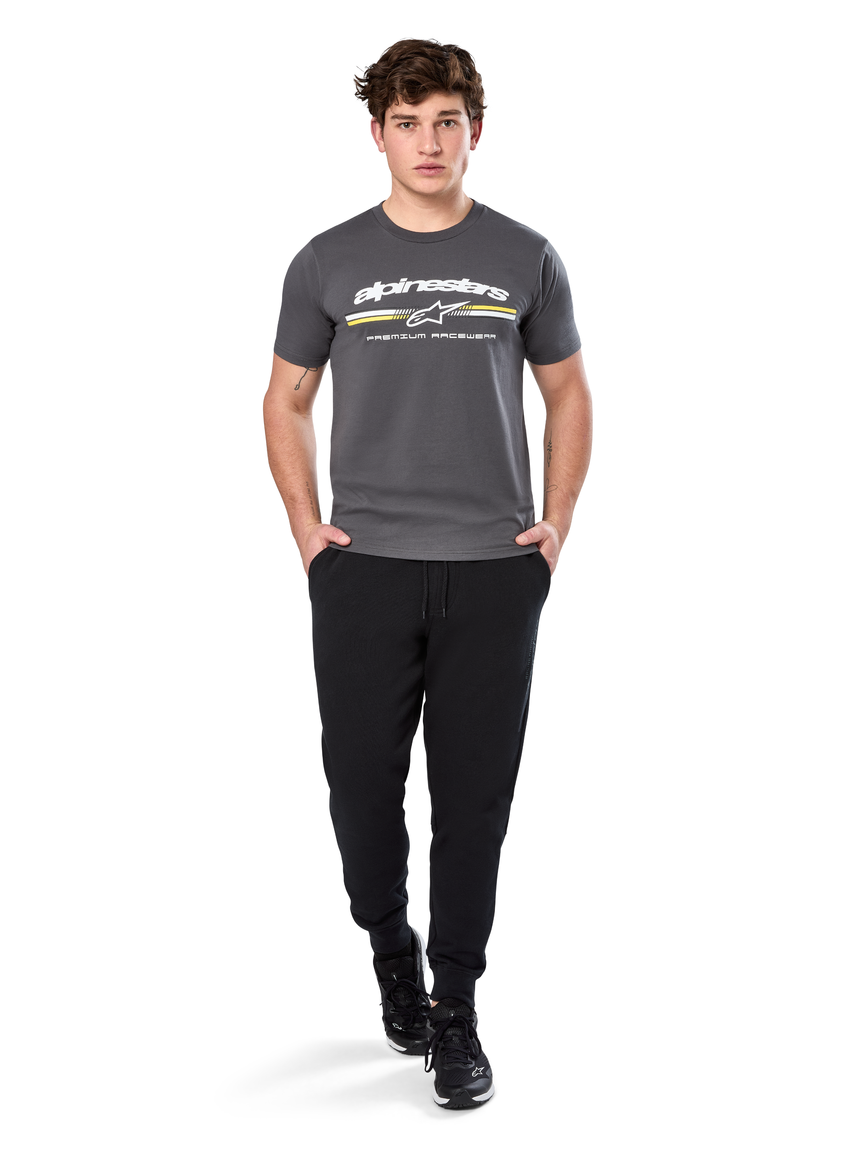 Homme portant le t-shirt à manches courtes Alpinestars Prevail CSF, gris anthracite, avec logo Alpinestars blanc et rayure tricolore aux accents jaunes, associé à un pantalon de jogging noir et des baskets, style motorsport décontracté