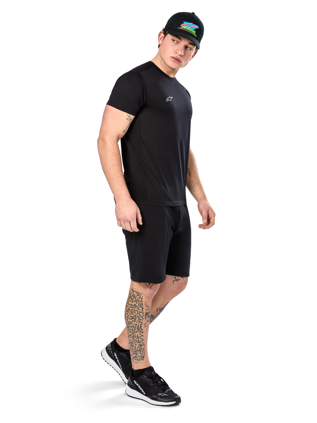 Man with tattoos wearing Alpinestars Point Performance Tee, Short sleeve black performance t-shirt, small white logo, paired with black shorts, a black cap with colorful graphic, and black and white sneakers, sporty casual lifestyle