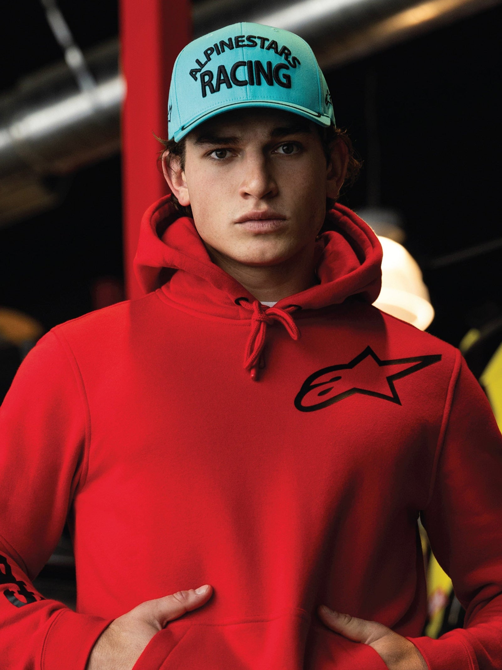 Casquette Luminary Alpinestars, snapback bleu aqua à visière incurvée, broderie 3D noire avec texte « ALPINESTARS RACING », portée avec un sweat à capuche Alpinestars rouge vif, style streetwear décontracté motorsport lifestyle