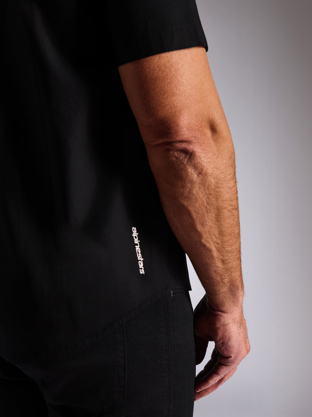 Chemise Alpinestars Strive Woven, chemise noire boutonnée à manches courtes, logo Alpinestars vertical blanc sur le côté, gros plan sur l'ourlet inférieur et le logo de la marque, style streetwear décontracté inspiré du sport automobile