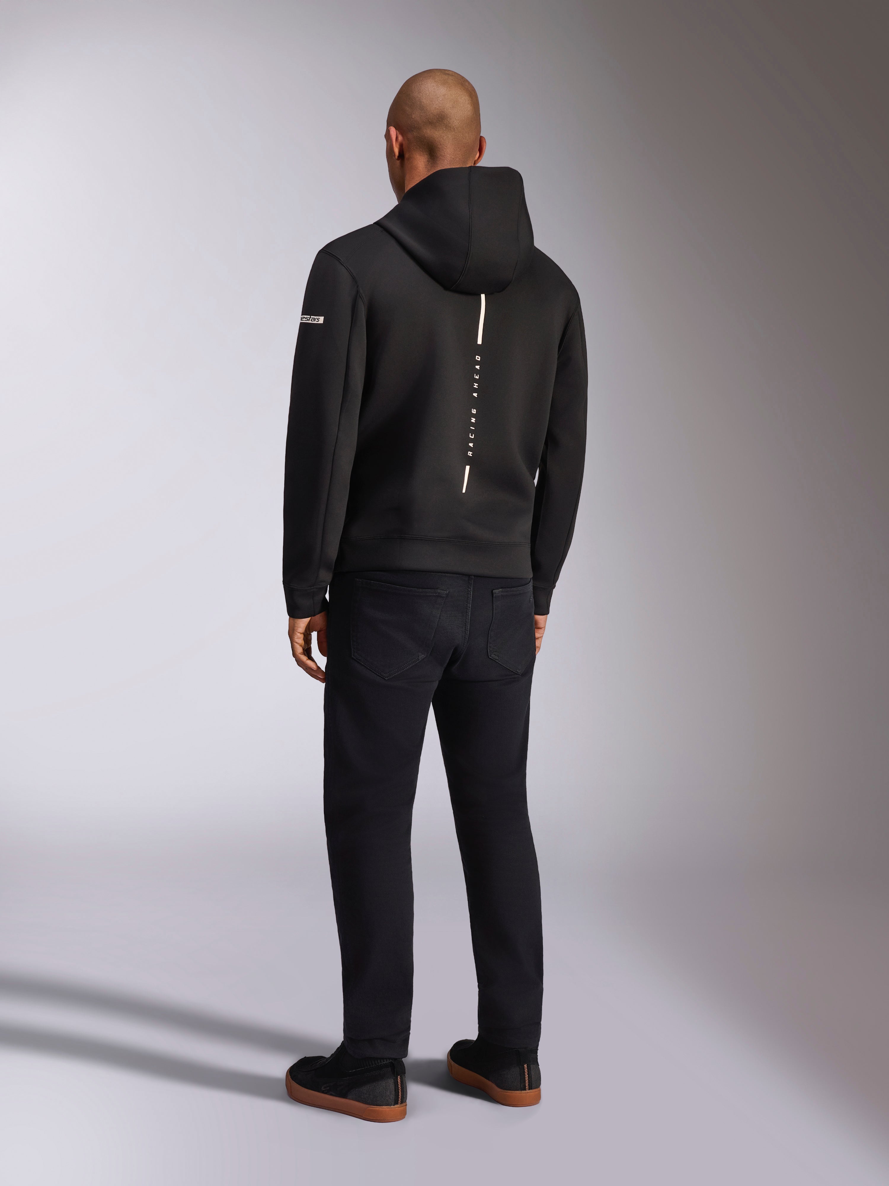 Alpinestars Insight Hoodie, Black sportswear hoodie, back view showing a vertical white stripe with 'RACING AHEAD' branding, sleeve logo, paired with black pants and sneakers, casual motorsport-inspired design
