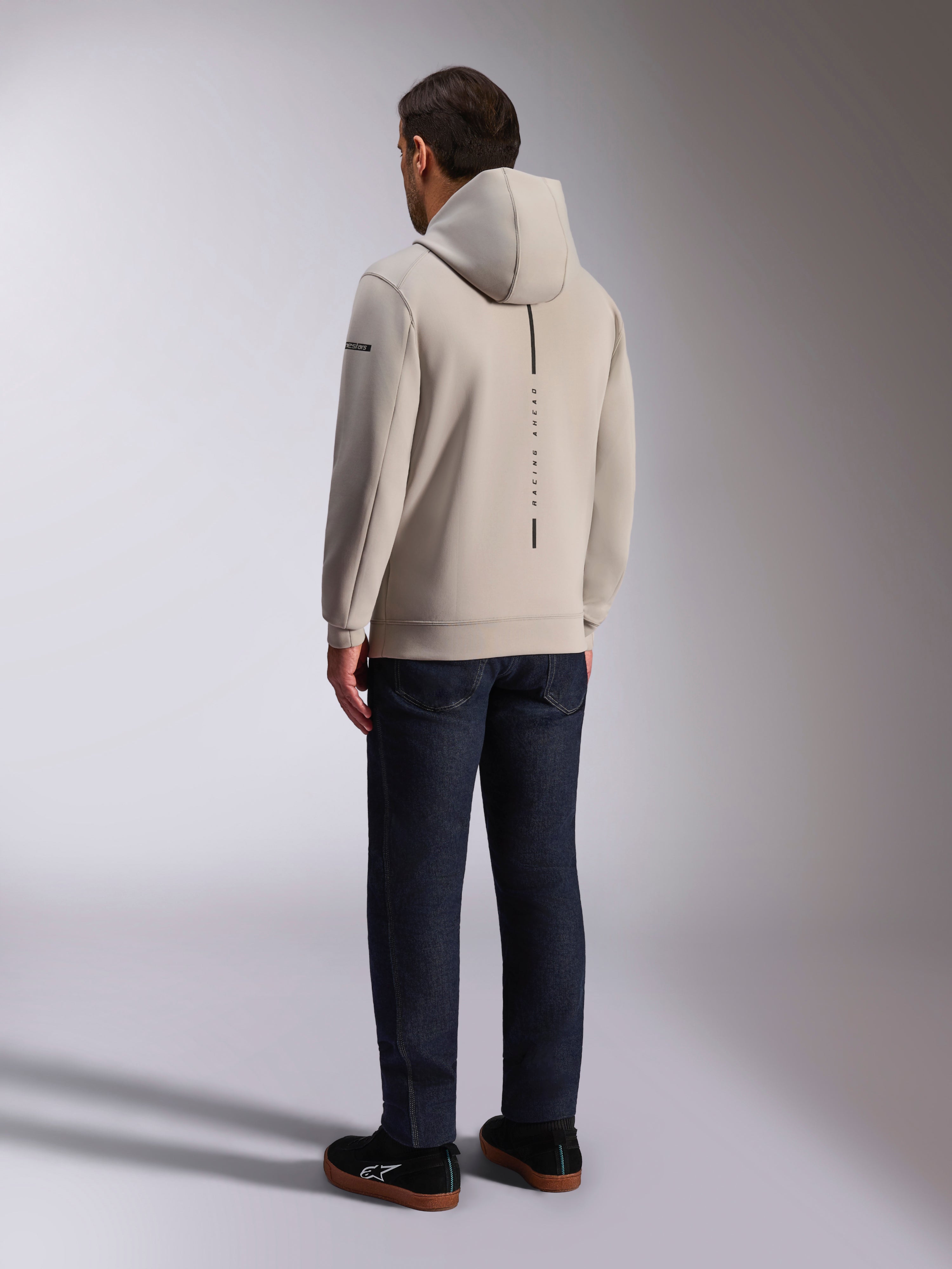 Sweat à capuche Alpinestars Insight, vue de dos en gris ciment, bande noire verticale avec le texte « RACING AHEAD », design streetwear décontracté, porté avec un jean foncé et des chaussures noires.