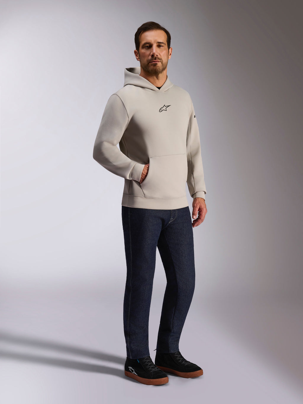 Sweat à capuche Insight Alpinestars, streetwear décontracté, gris ciment avec logo Astars noir sur la poitrine, design à enfiler avec poche kangourou, porté avec un jean bleu foncé et des baskets noires, style moderne inspiré du sport automobile