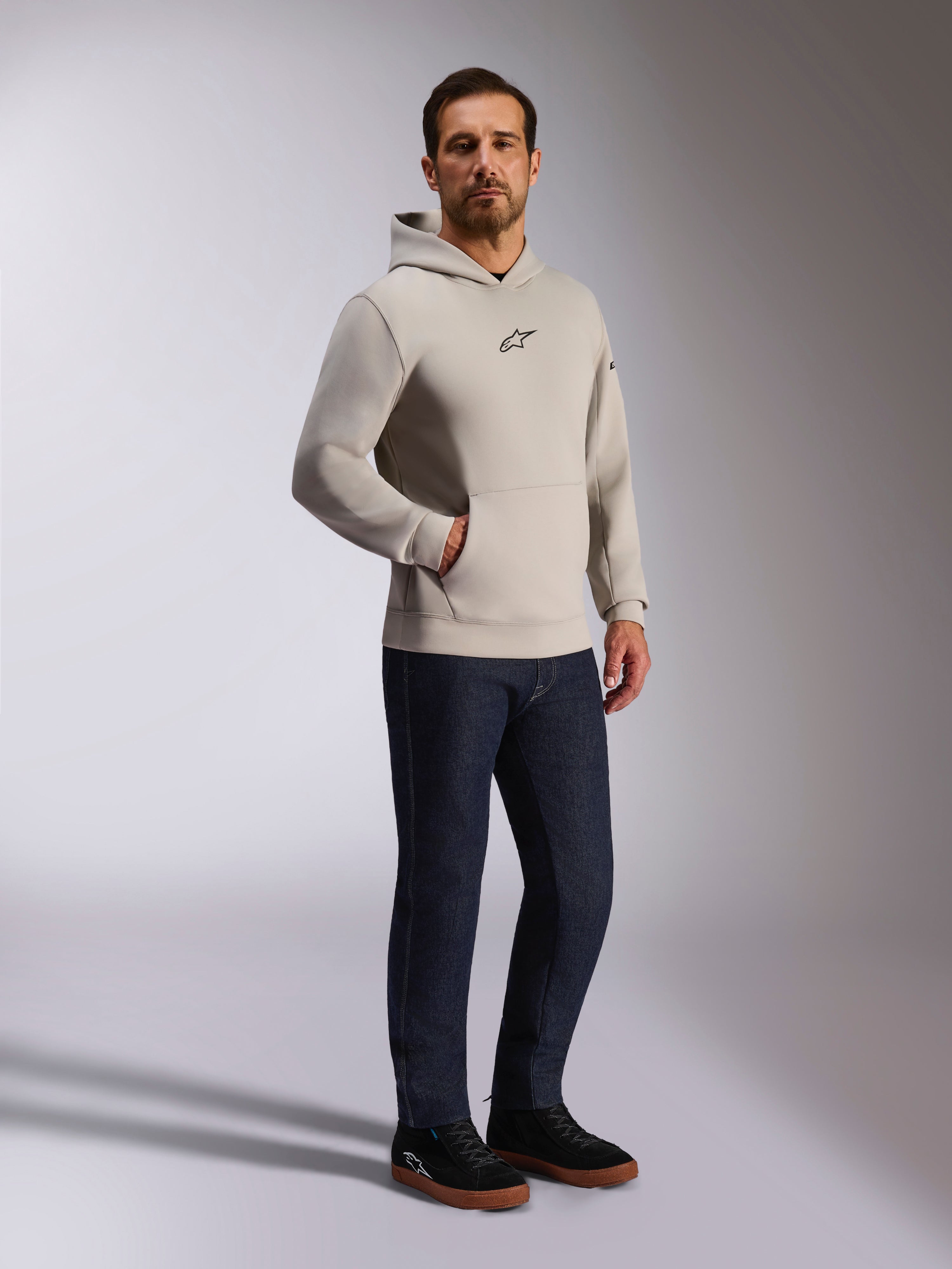 Sweat à capuche Insight Alpinestars, streetwear décontracté, gris ciment avec logo Astars noir sur la poitrine, design à enfiler avec poche kangourou, porté avec un jean bleu foncé et des baskets noires, style moderne inspiré du sport automobile