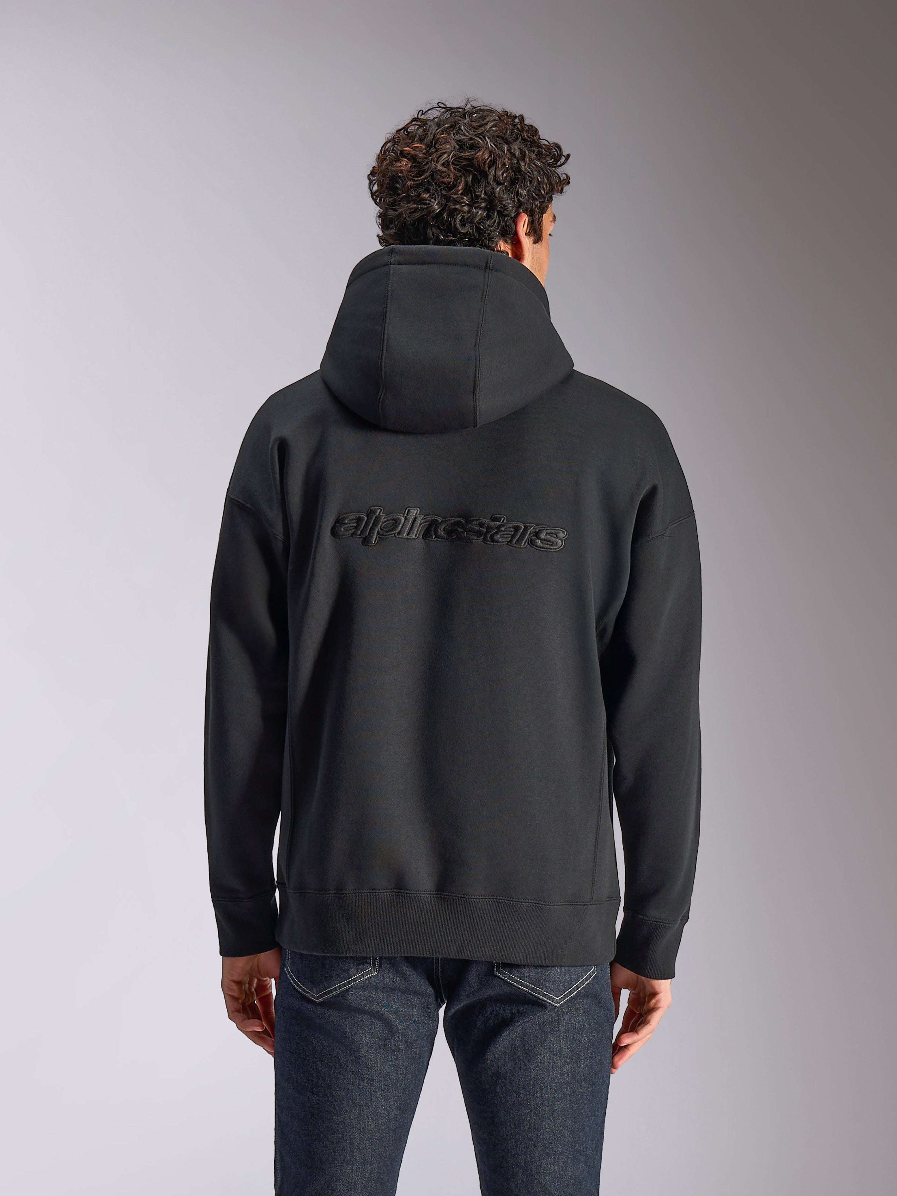 Sweat à capuche Alpinestars Emergent Oversized, vue de dos d'un sweat à capuche noir sur un mannequin, logo Alpinestars brodé au dos, coupe ample, style streetwear décontracté associé à un jean foncé