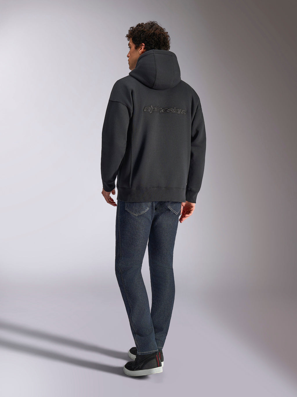 Sweat à capuche Alpinestars Emergent Oversized, noir, vue de dos montrant le logo Alpinestars en broderie 3D sur les épaules, coupe ample, porté avec un jean en denim bleu foncé et des baskets noires, streetwear décontracté inspiré du sport automobile.