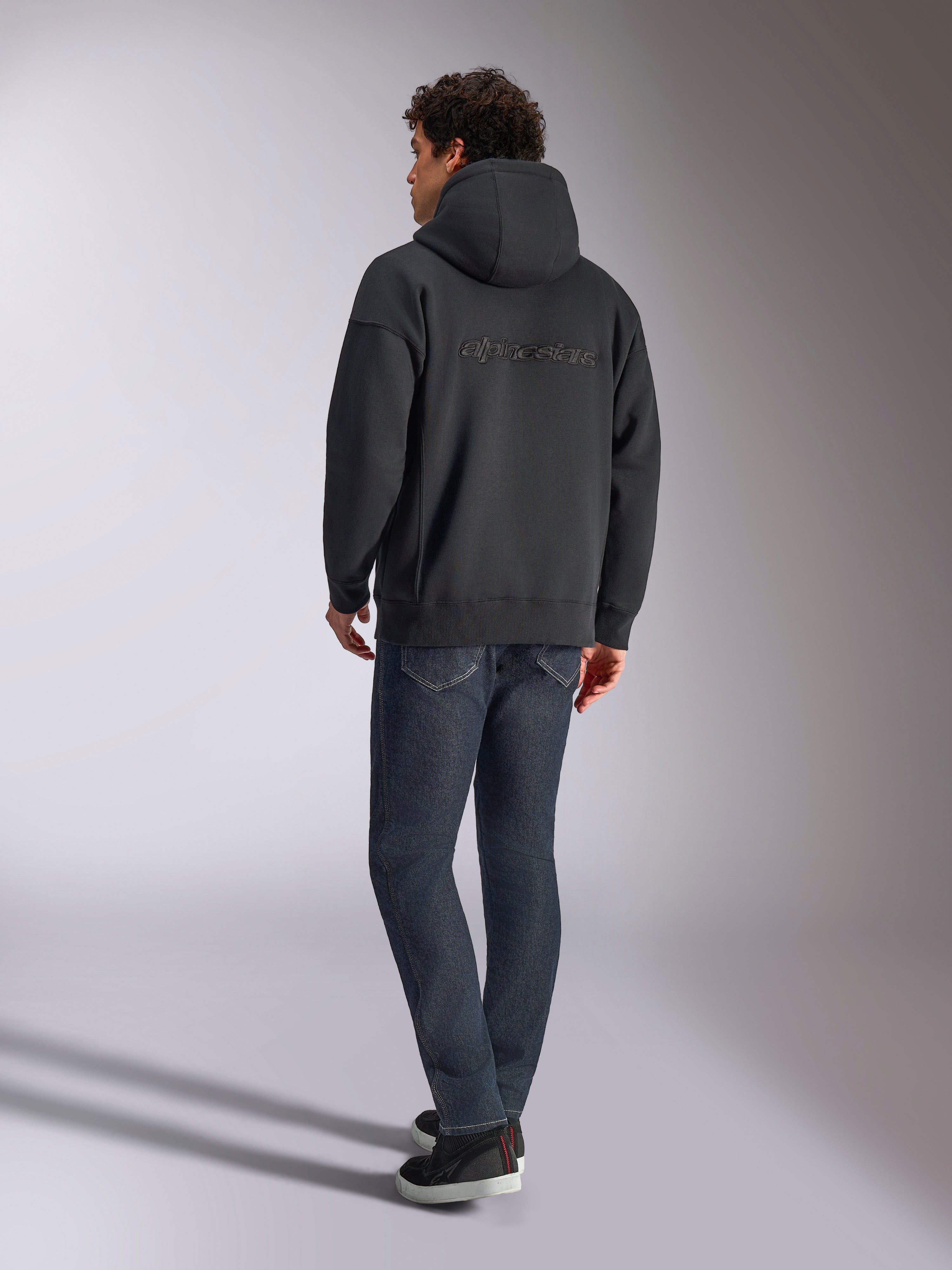 Sweat à capuche Alpinestars Emergent Oversized, noir, vue de dos montrant le logo Alpinestars en broderie 3D sur les épaules, coupe ample, porté avec un jean en denim bleu foncé et des baskets noires, streetwear décontracté inspiré du sport automobile.