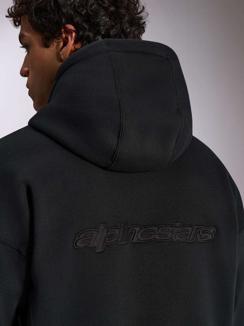 Sweat à capuche Alpinestars Emergent Oversized, vêtement streetwear décontracté noir, vue de dos avec logo Alpinestars en lettres brodées 3D, coupe ample et texture polaire premium