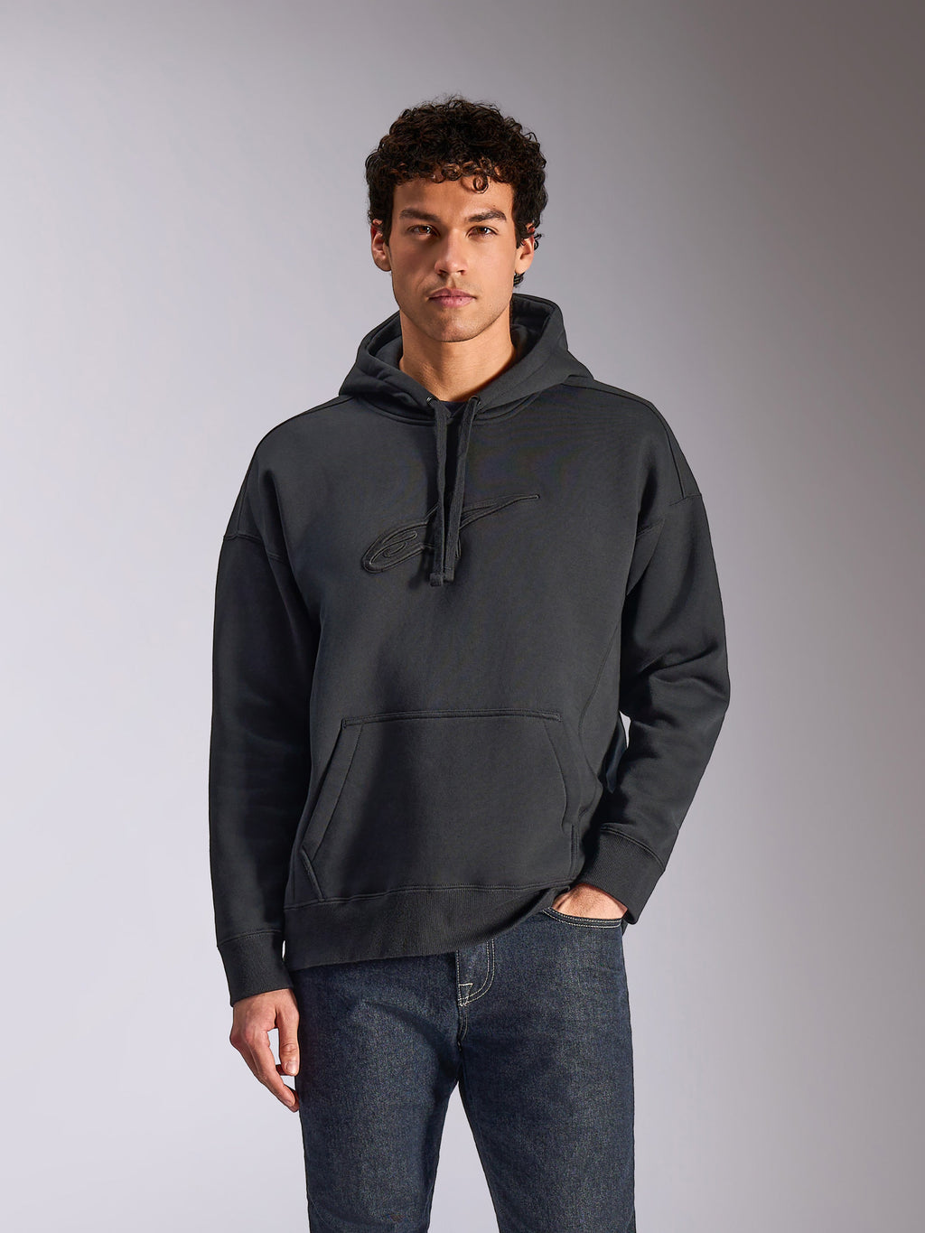Alpinestars Emergent Oversized Hoodie, noir, mannequin masculin portant un sweat à capuche en polaire de coupe ample, logo Astars brodé en 3D ton sur ton, poche kangourou, capuche à cordon de serrage, streetwear décontracté inspiré du sport automobile