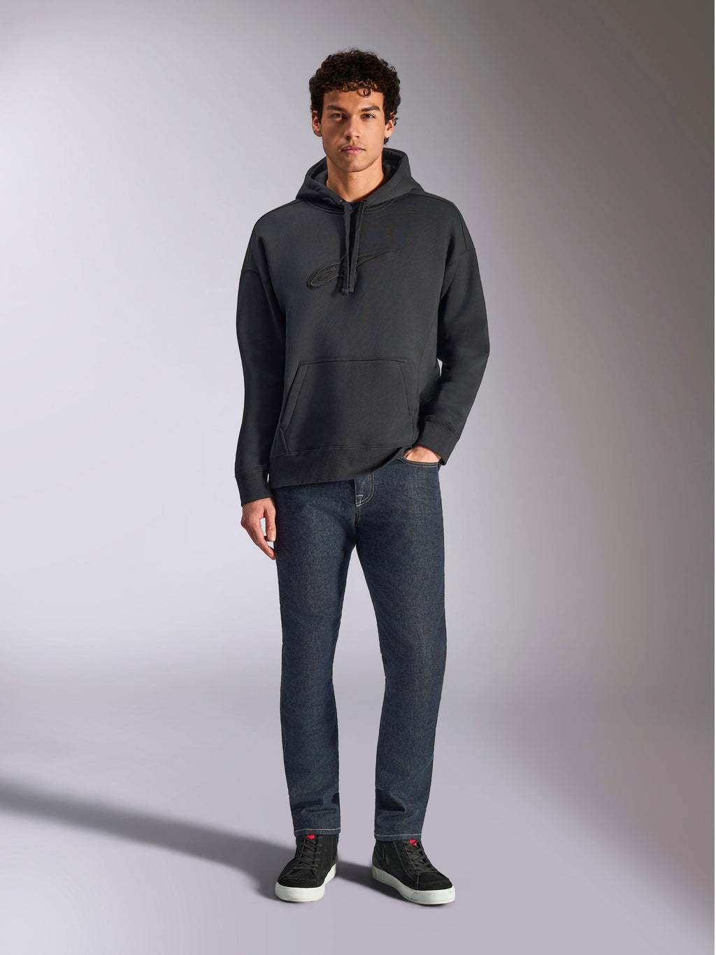 Alpinestars Emergent Oversized Hoodie, sweat à capuche de style streetwear décontracté, noir avec logo brodé en 3D ton sur ton, capuche à cordon de serrage, poche kangourou, coupe ample, porté par un mannequin avec un jean bleu foncé et des baskets noires