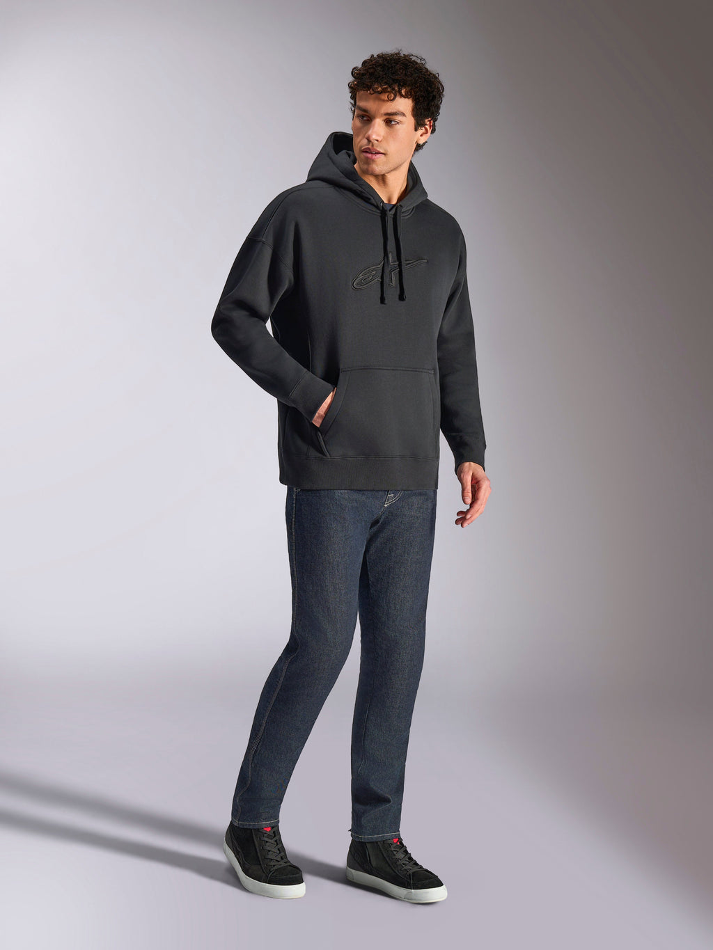 Sweat à capuche oversized Emergent Alpinestars, streetwear décontracté noir, logo Astars brodé en 3D ton sur ton sur la poitrine, poche kangourou, coupe oversized, porté par un mannequin homme avec un jean en denim bleu foncé et des baskets noires