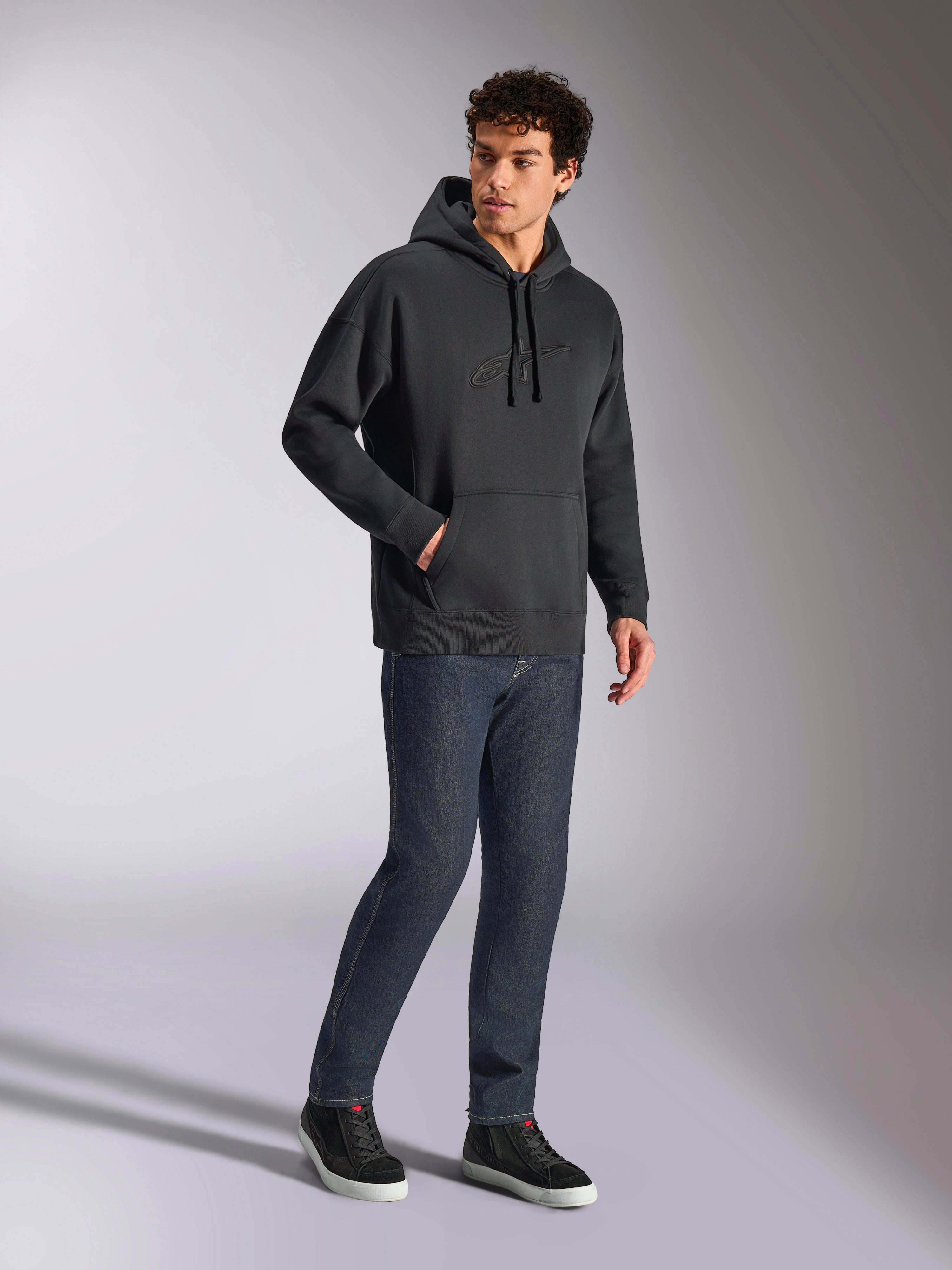 Sweat à capuche oversized Emergent Alpinestars, streetwear décontracté noir, logo Astars brodé en 3D ton sur ton sur la poitrine, poche kangourou, coupe oversized, porté par un mannequin homme avec un jean en denim bleu foncé et des baskets noires