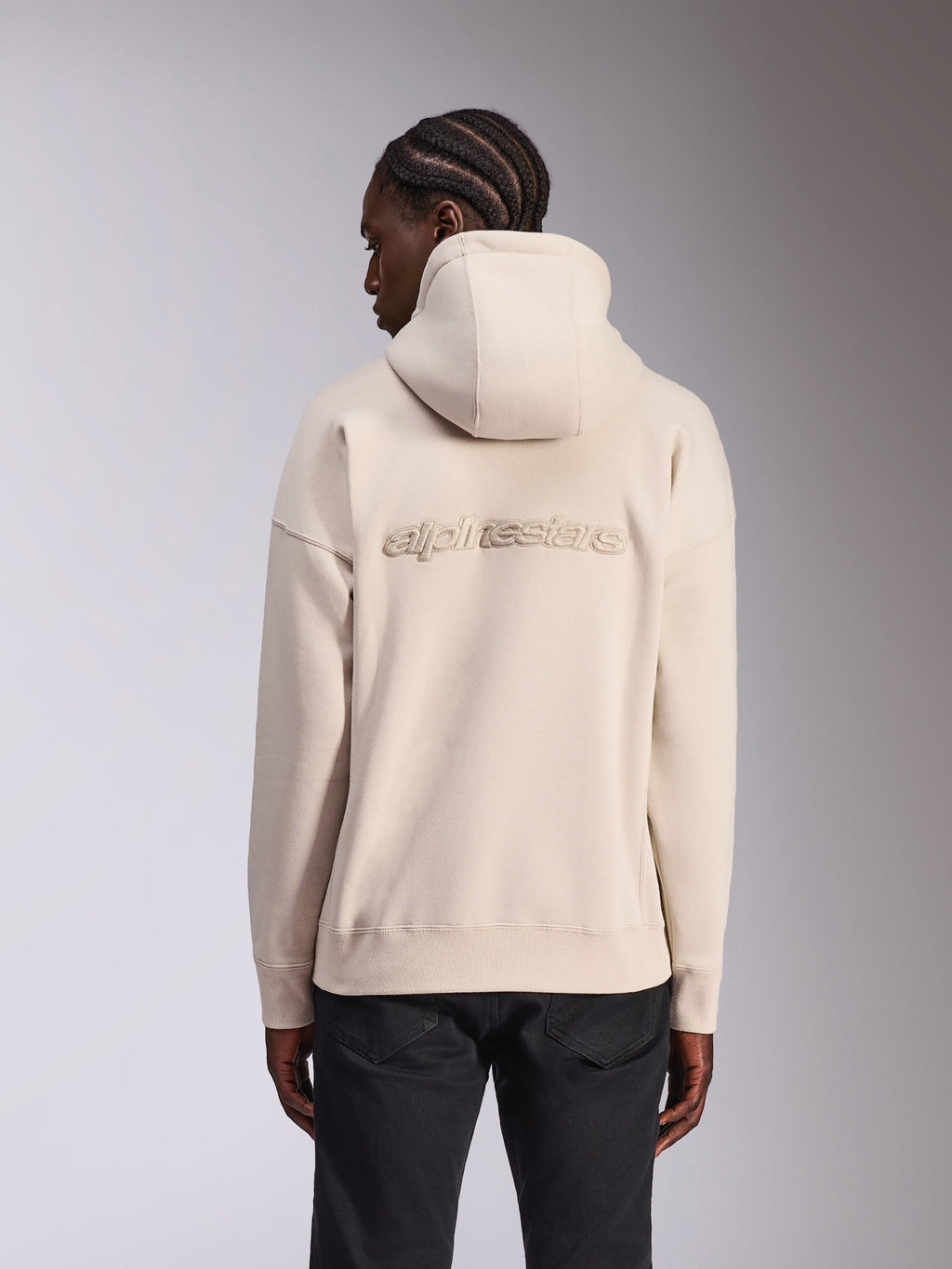 Sweat à capuche oversized Emergent Alpinestars, streetwear décontracté, couleur sable blanc cassé, avec logo Alpinestars brodé en 3D dans le dos, coupe ample avec ourlet et poignets côtelés, vue de dos sur mannequin portant un pantalon noir.