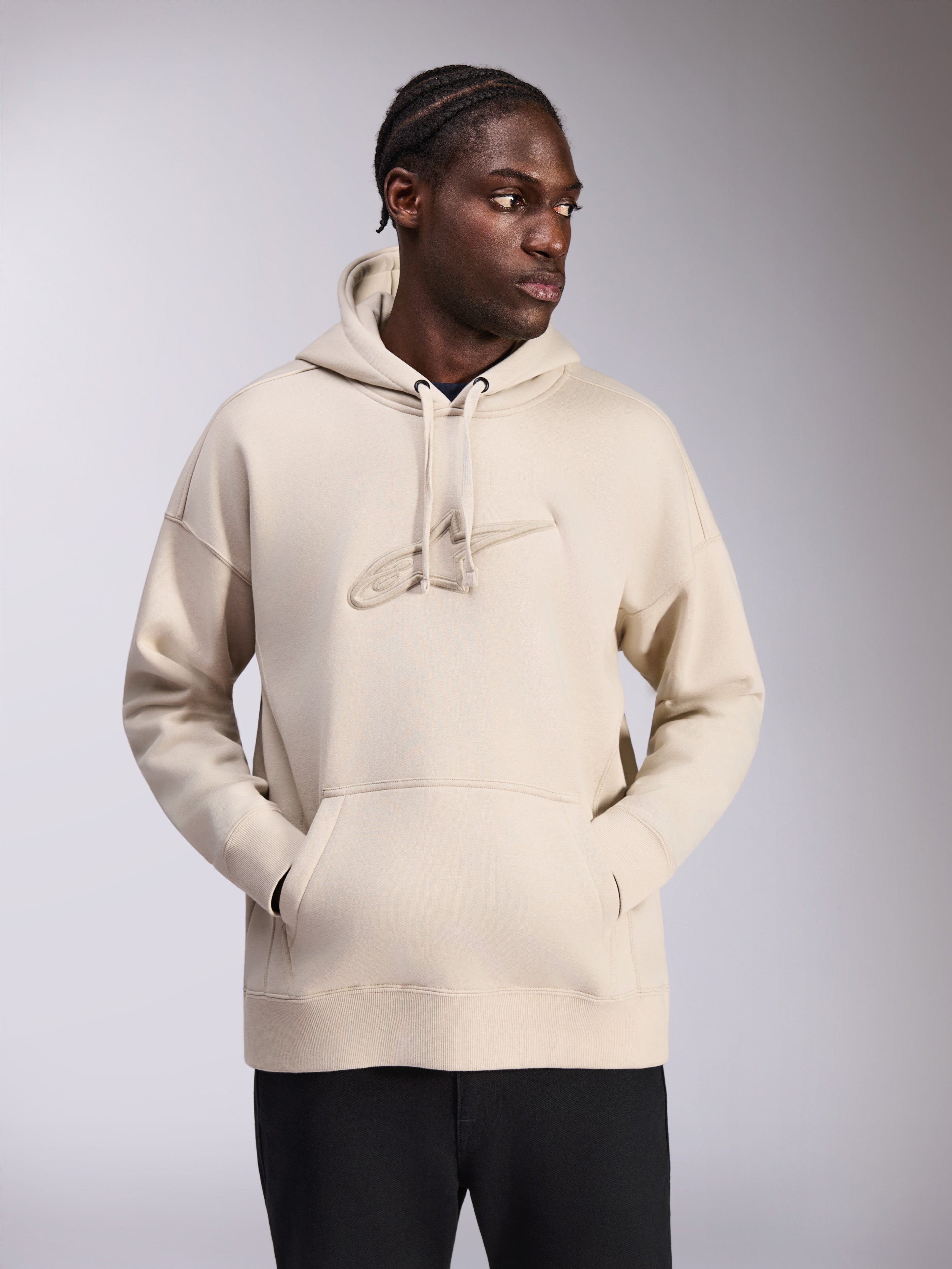Sweat à capuche oversize Emergent Alpinestars, couleur sable, coupe ample avec logo Astars brodé en 3D ton sur ton sur la poitrine, poche kangourou, capuche à cordon de serrage, style streetwear décontracté porté par un mannequin avec un pantalon noir