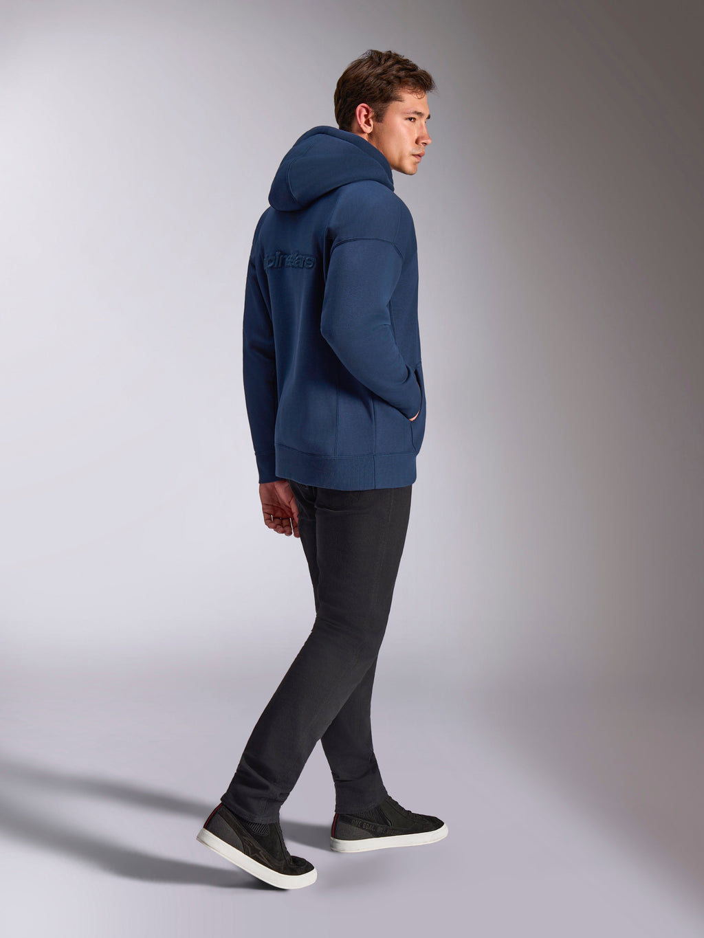 Sweat à capuche oversized Alpinestars Emergent bleu marine, vue de dos montrant le logo Alpinestars brodé en 3D, coupe ample en polaire avec capuche à cordon de serrage, porté avec un pantalon noir, style streetwear décontracté inspiré du sport automobile