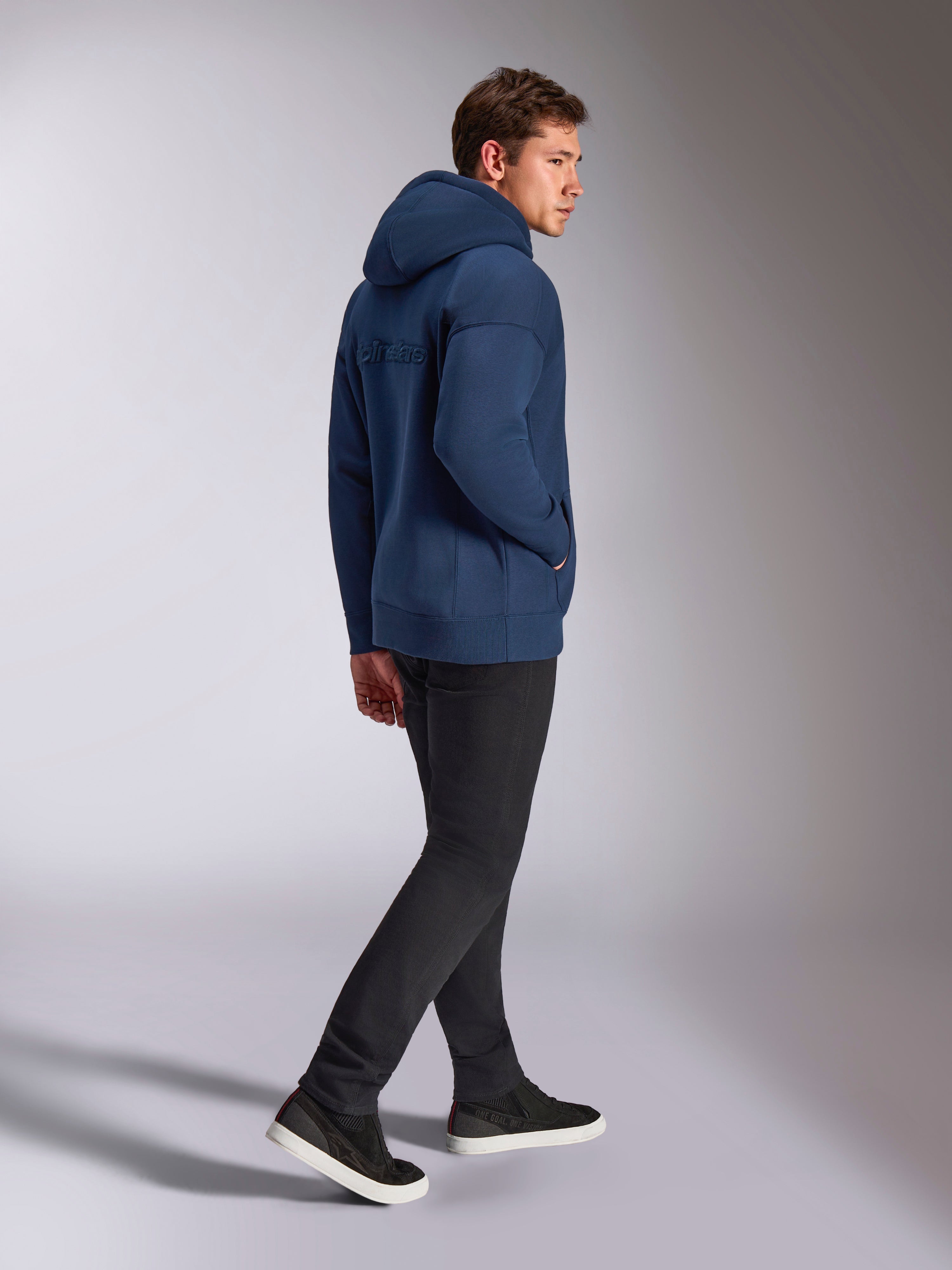 Sweat à capuche oversized Alpinestars Emergent bleu marine, vue de dos montrant le logo Alpinestars brodé en 3D, coupe ample en polaire avec capuche à cordon de serrage, porté avec un pantalon noir, style streetwear décontracté inspiré du sport automobile