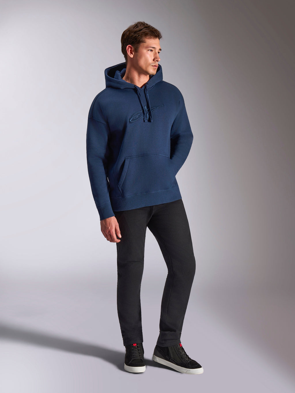 Sweat à capuche oversized Emergent Alpinestars, bleu marine, porté par un mannequin homme avec un pantalon noir et des baskets, avec logo Astars brodé en 3D, poche kangourou et capuche à cordon de serrage, style streetwear décontracté inspiré du sport automobile.