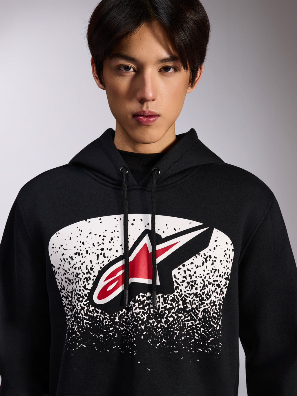 Alpinestars Obsessed Hoodie, sweat à capuche en polaire noire, logo Astars rouge et blanc dans un graphisme en dégradé MX pointillé blanc sur la poitrine, porté par un mannequin, streetwear décontracté avec capuche à cordon de serrage ajustable