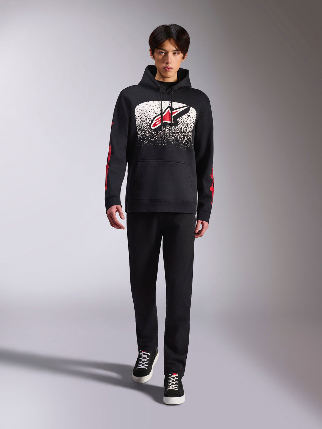Sweat à capuche Alpinestars Obsessed, sweat à capuche noir à enfiler, grand logo Astars blanc et rouge avec dégradé MX, logos Alpinestars Word rouges sur les manches, porté par un mannequin dans une tenue décontractée entièrement noire, vêtement lifestyle inspiré du sport automobile