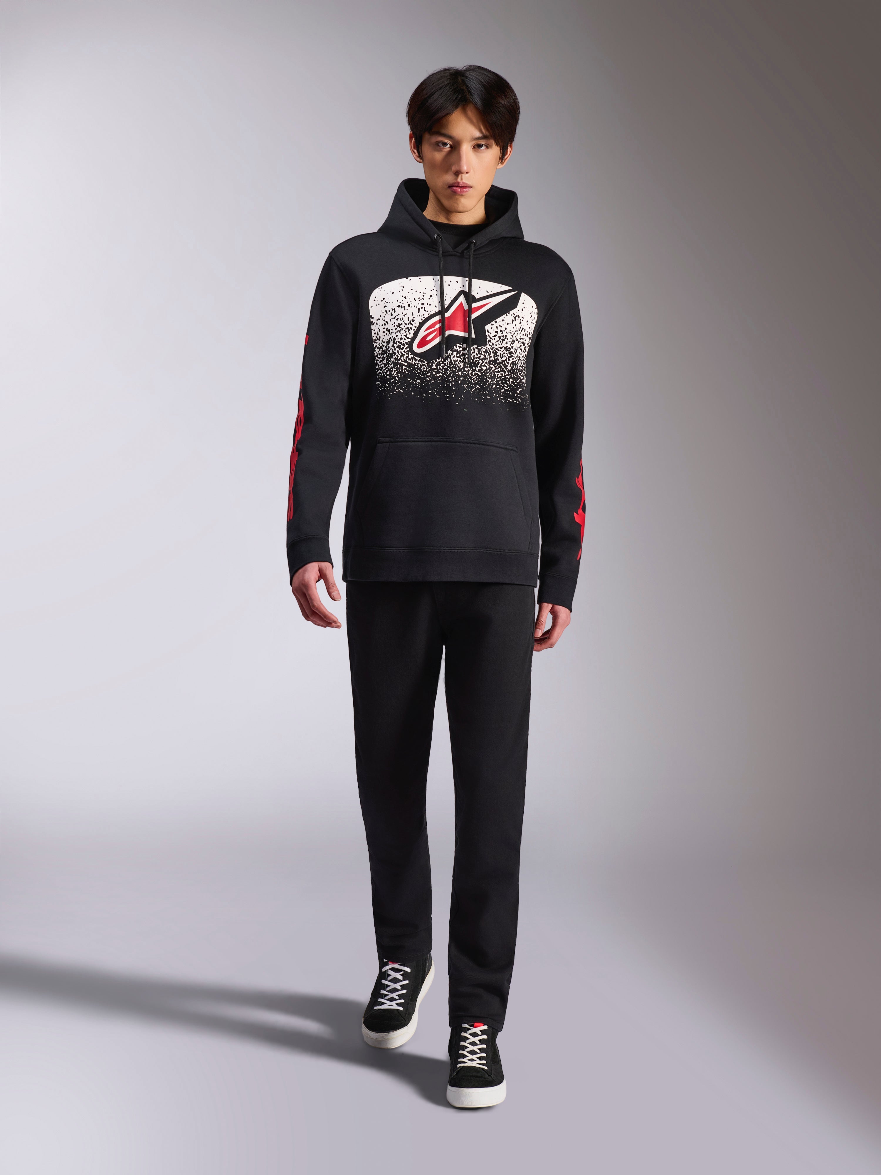 Sweat à capuche Alpinestars Obsessed, sweat à capuche noir à enfiler, grand logo Astars blanc et rouge avec dégradé MX, logos Alpinestars Word rouges sur les manches, porté par un mannequin dans une tenue décontractée entièrement noire, vêtement lifestyle inspiré du sport automobile