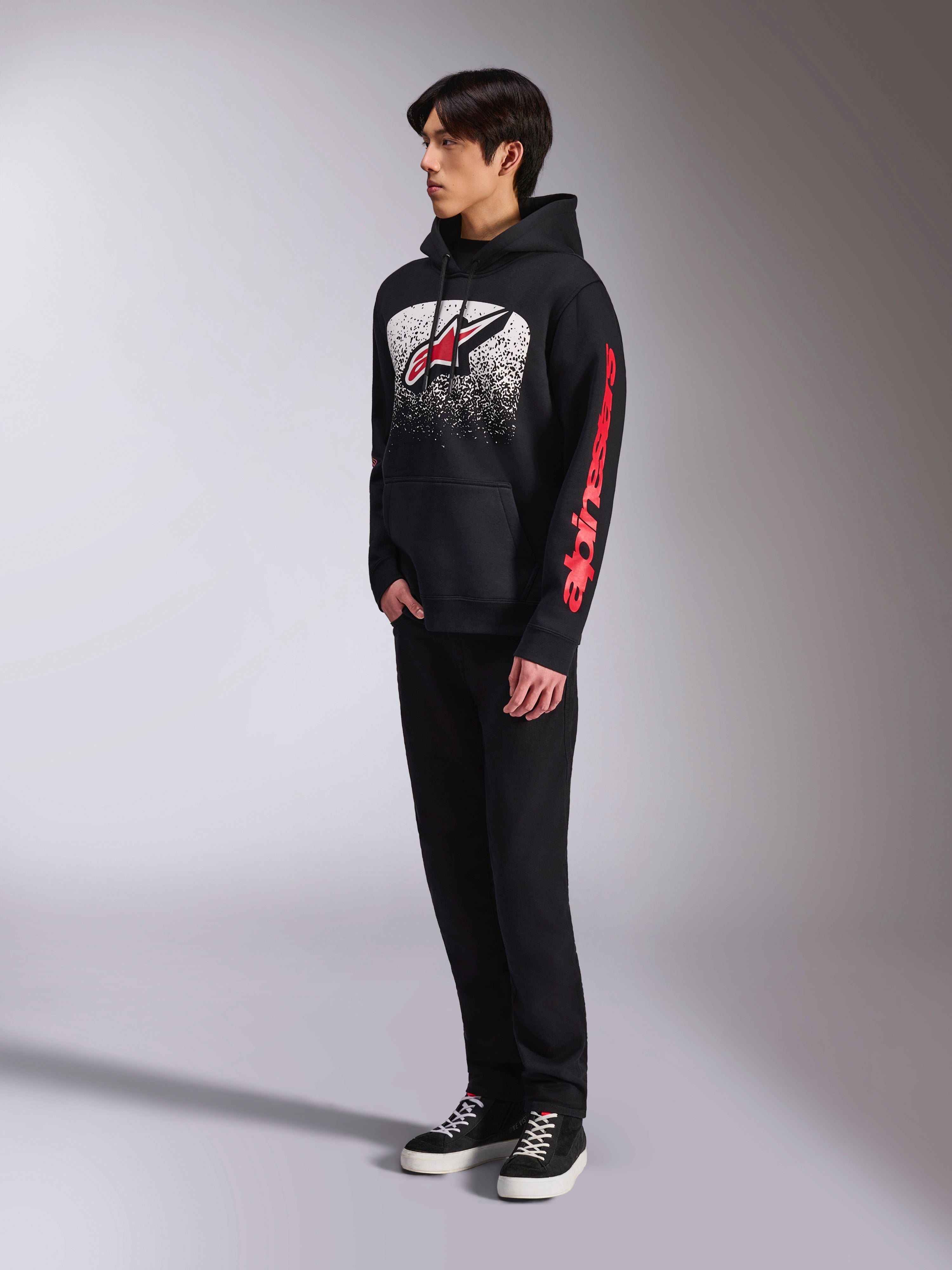 Sweat à capuche Alpinestars Obsessed, sweat en polaire noir à enfiler, avec un grand graphisme blanc moucheté sur la poitrine, logo Astars rouge et marquage rouge sur la manche, porté par un mannequin avec pantalon noir et baskets, style streetwear motorsport décontracté