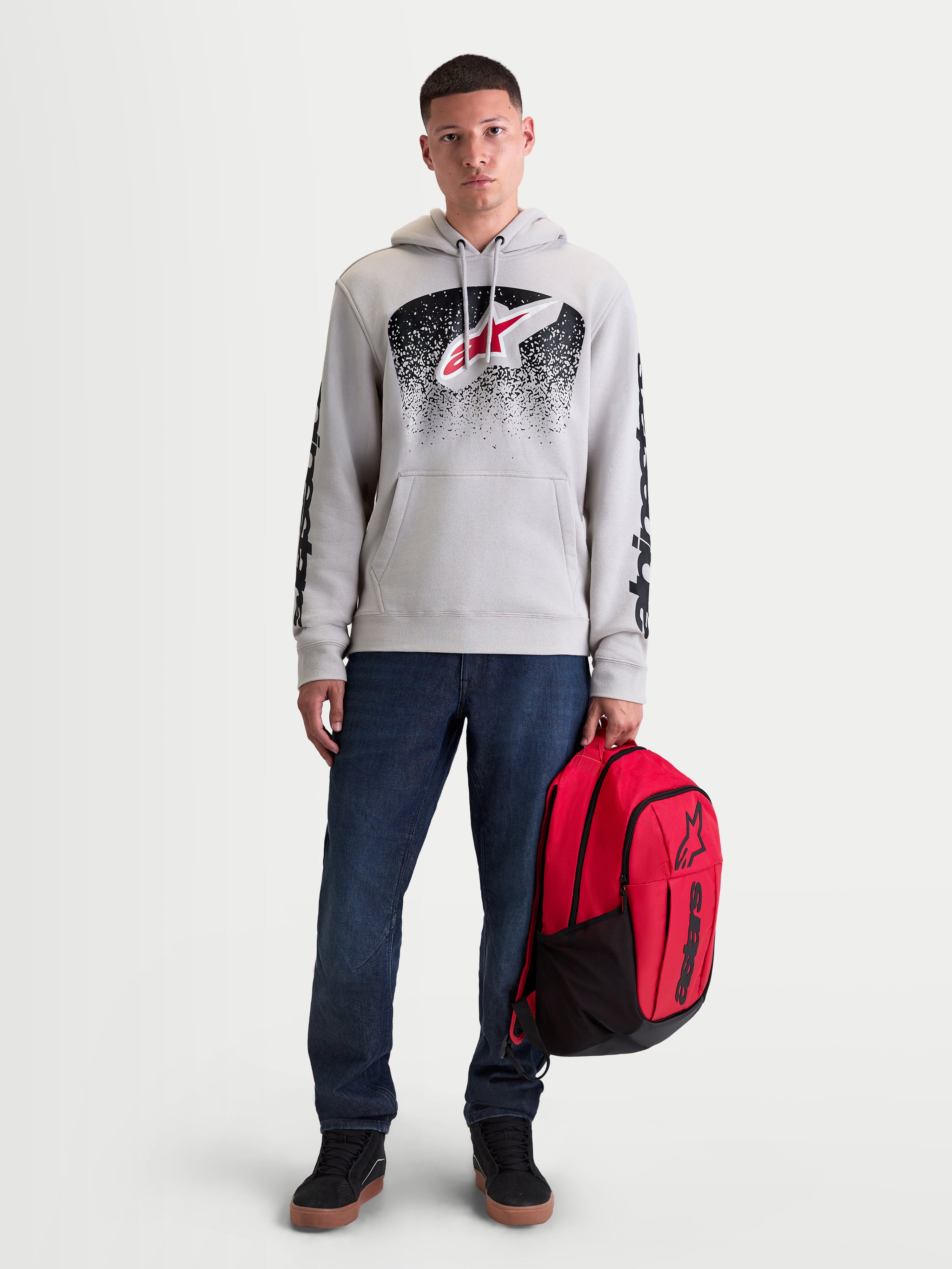 Alpinestars Sweat À Capuche Obsessed, sweat de sportswear en polaire pour homme, gris ciment avec graphismes dégradés rouges et noirs d'inspiration MX, logos sur les manches, porté par un modèle tenant un sac à dos Alpinestars rouge.