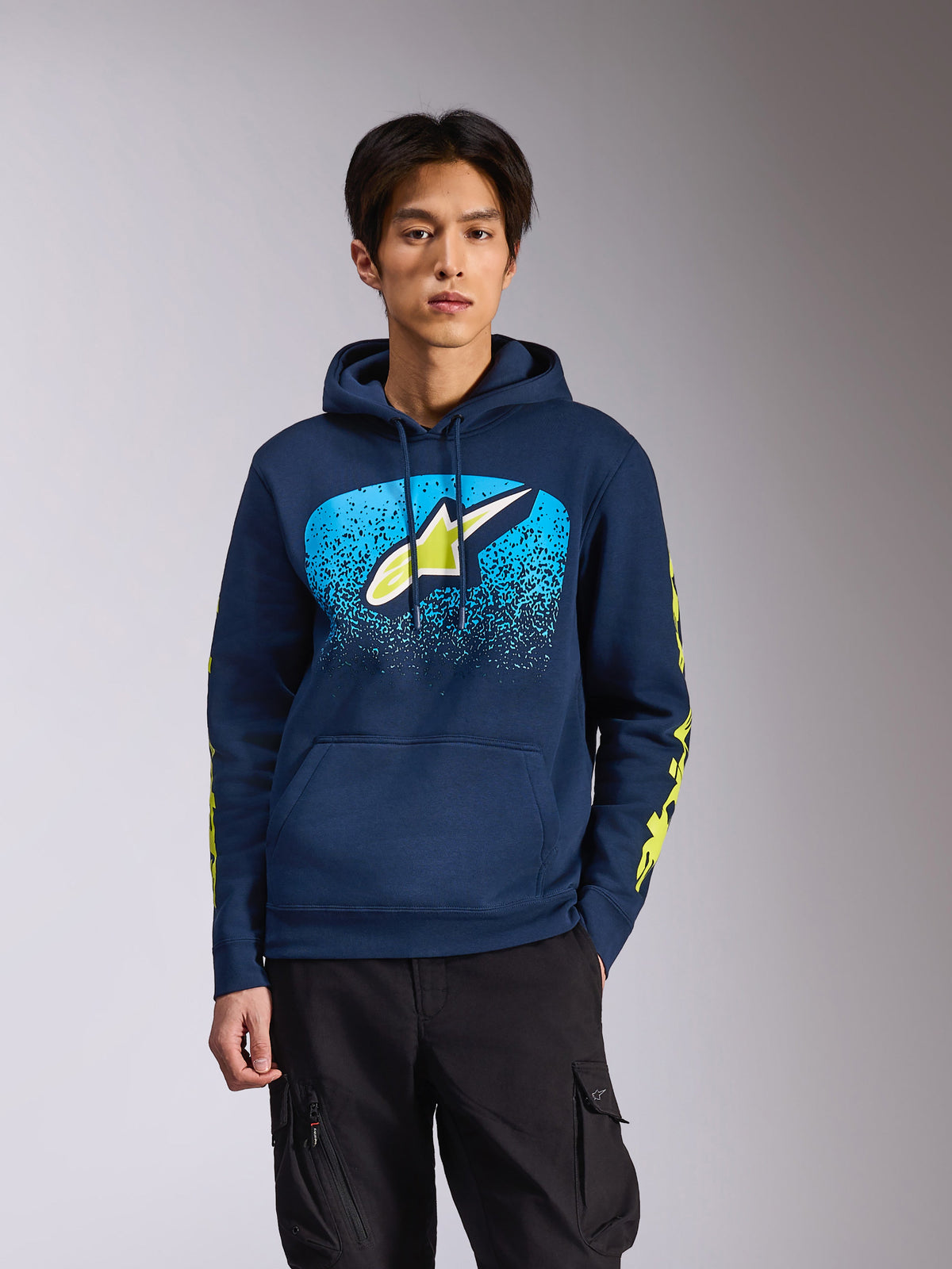 Sweat à capuche Alpinestars Obsessed bleu marine, porté par un mannequin homme, logo Astars vert citron avec dégradé MX bleu vif sur la poitrine, logos Alpinestars jaunes sur les manches, streetwear décontracté inspiré du sport mécanique