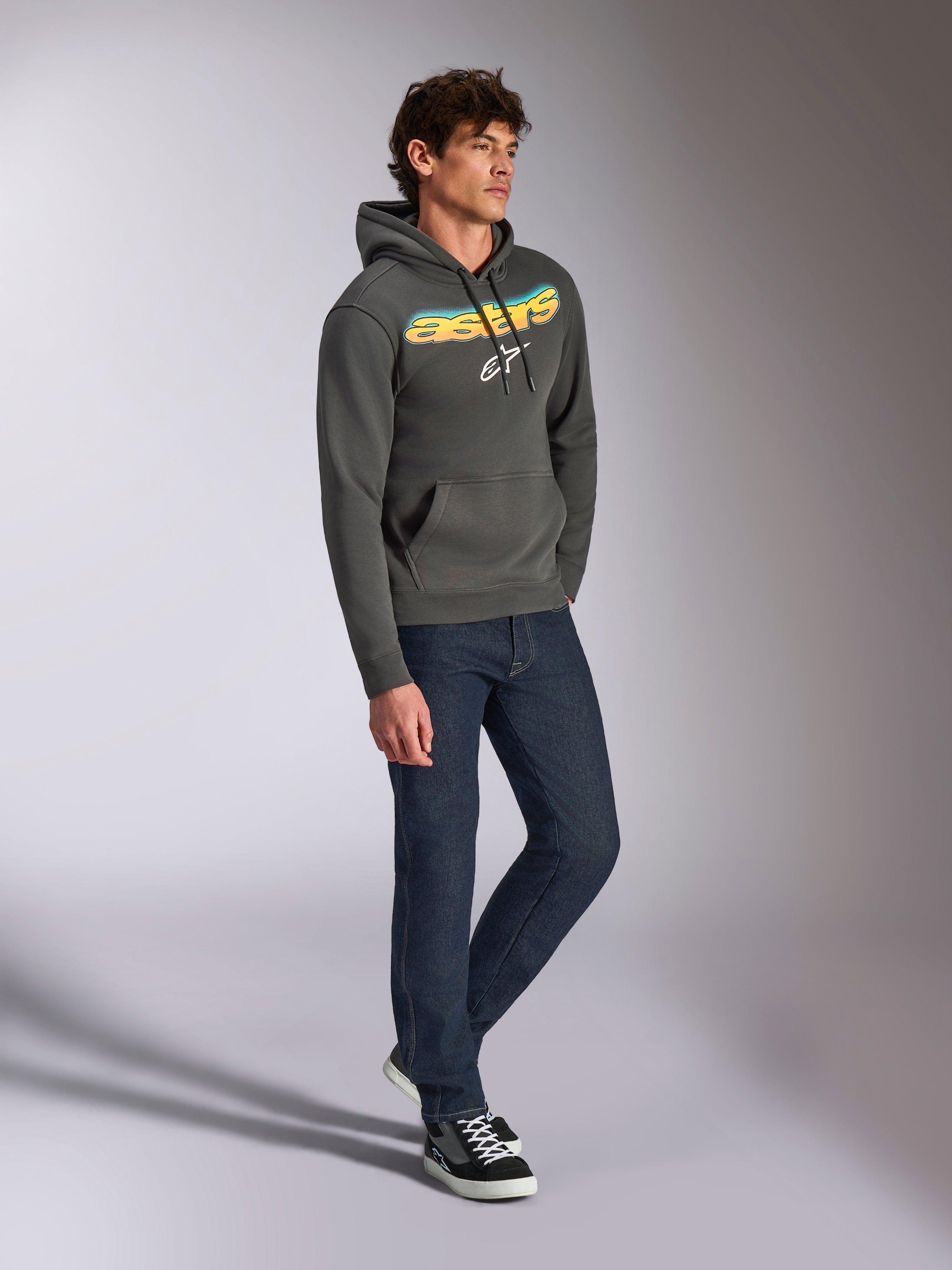 Sweat à capuche Alpinestars Ellipsoid, polaire à enfiler gris anthracite, lettrage Astars incurvé à l'avant avec dégradé bleu et jaune et logo blanc, porté avec un jean foncé et des baskets noires, style streetwear décontracté.