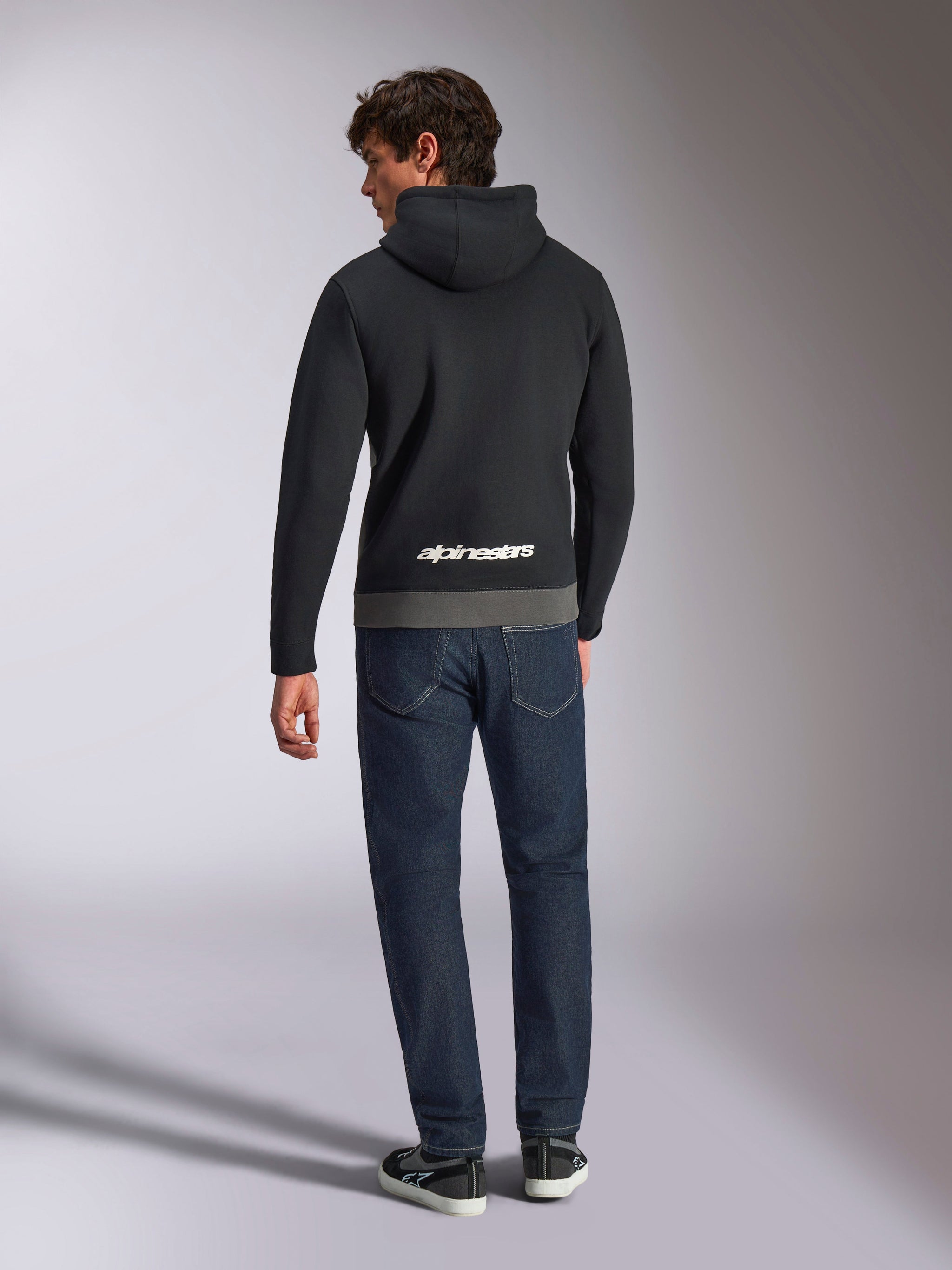 Sweat à capuche Parallel Alpinestars, vue de dos d'un homme portant un sweat en polaire à enfiler noir et anthracite, logo Alpinestars blanc dans le bas du dos, associé à un jean bleu foncé, style streetwear décontracté inspiré des sports mécaniques