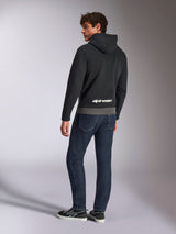 Sweat à capuche Parallel d'Alpinestars, vue arrière d'un mannequin portant le pullover noir et gris gunmetal, arborant le logo Alpinestars blanc sur le bas du dos, associé à un jean bleu foncé, style streetwear décontracté inspiré du motorsport