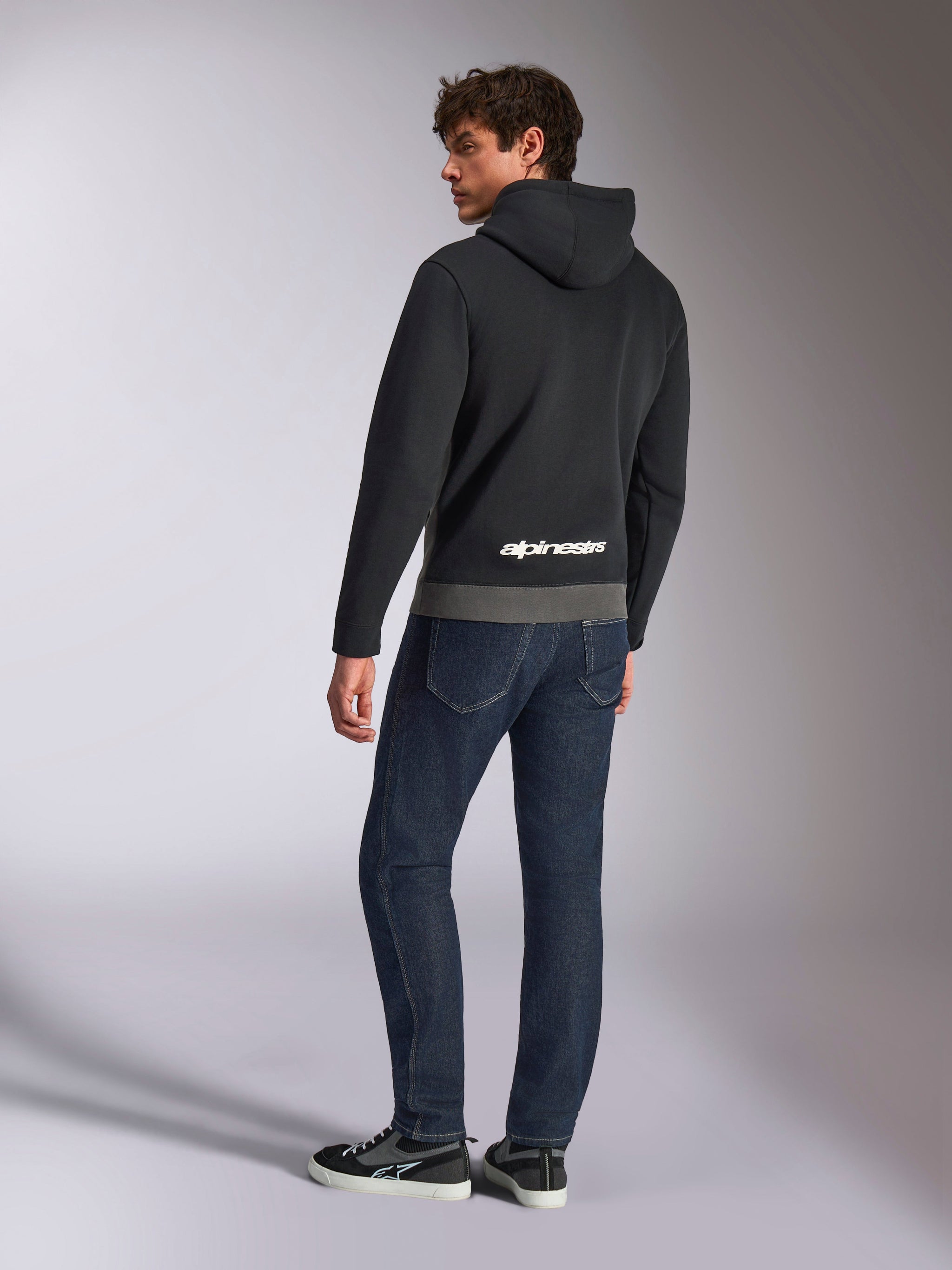 Sweat à capuche Parallel d'Alpinestars, vue arrière d'un mannequin portant le pullover noir et gris gunmetal, arborant le logo Alpinestars blanc sur le bas du dos, associé à un jean bleu foncé, style streetwear décontracté inspiré du motorsport